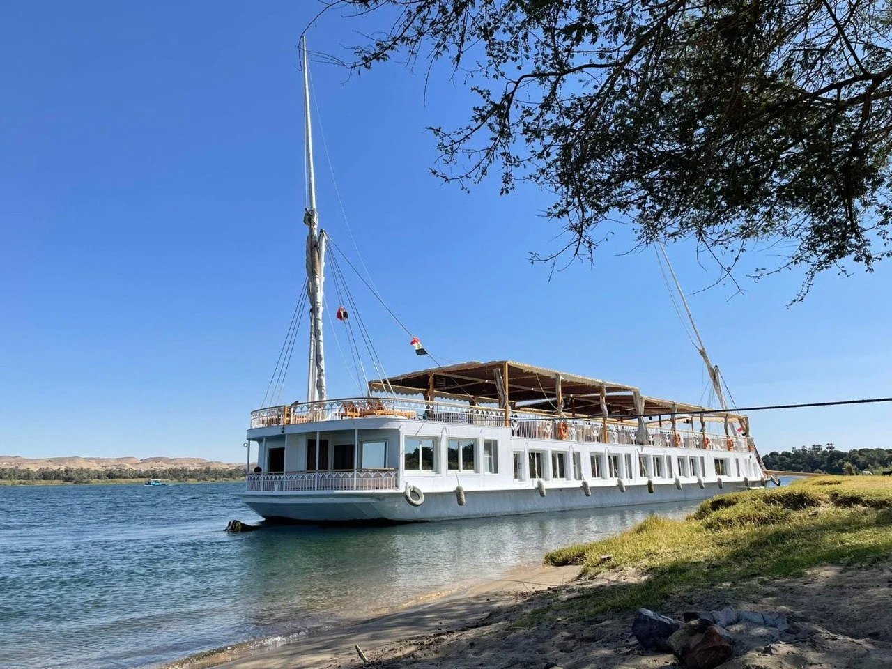 Ein weißes Binnenkreuzschiff mit offenen Oberdeck liegt am Ufer eines Flusses, umgeben von Gras und Bäumen bei schönem Himmel.