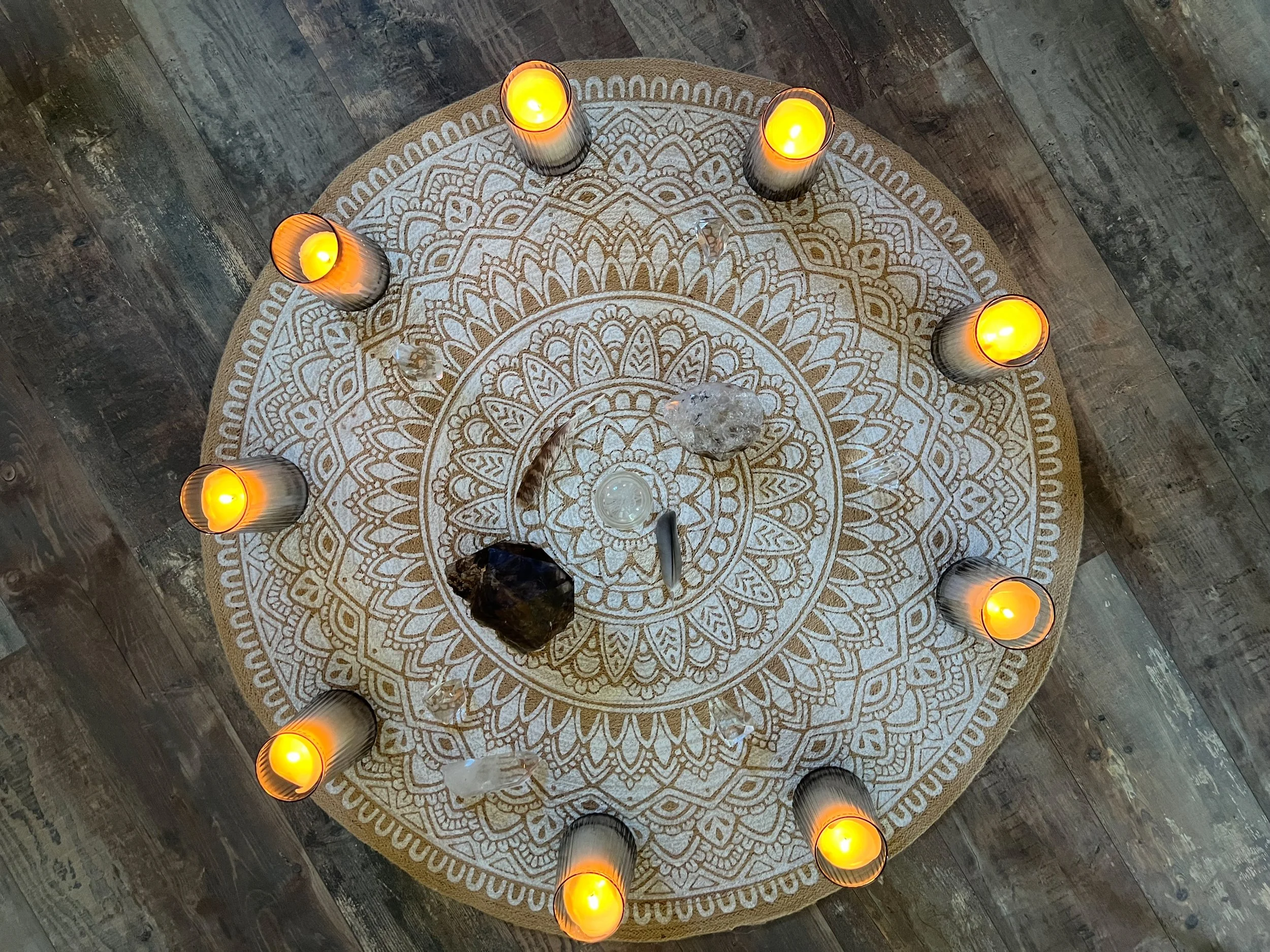 Runder Teppich mit weißen Mandala-Mustern, darauf sieben brennende Kerzen in schwarzen Kerzenhaltern, sowie Kristalle und eine kleine weiße Schale in der Mitte, auf einem Holzboden