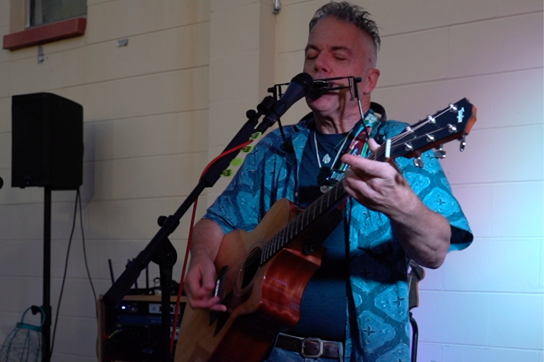 A man plays an acoustic guitar and sings into a microphone during a performance. He wears a blue patterned shirt and has gray hair.