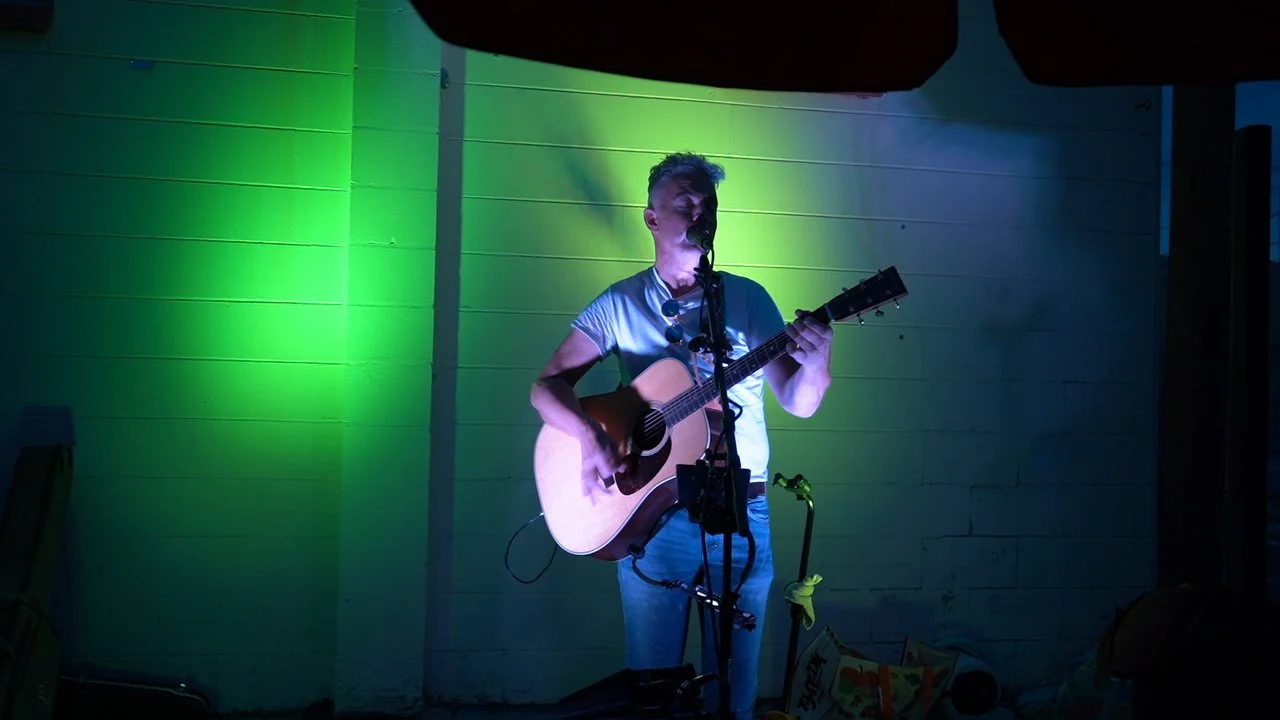 A young man plays an acoustic guitar and sings into a microphone on a stage illuminated with green and purple lights.