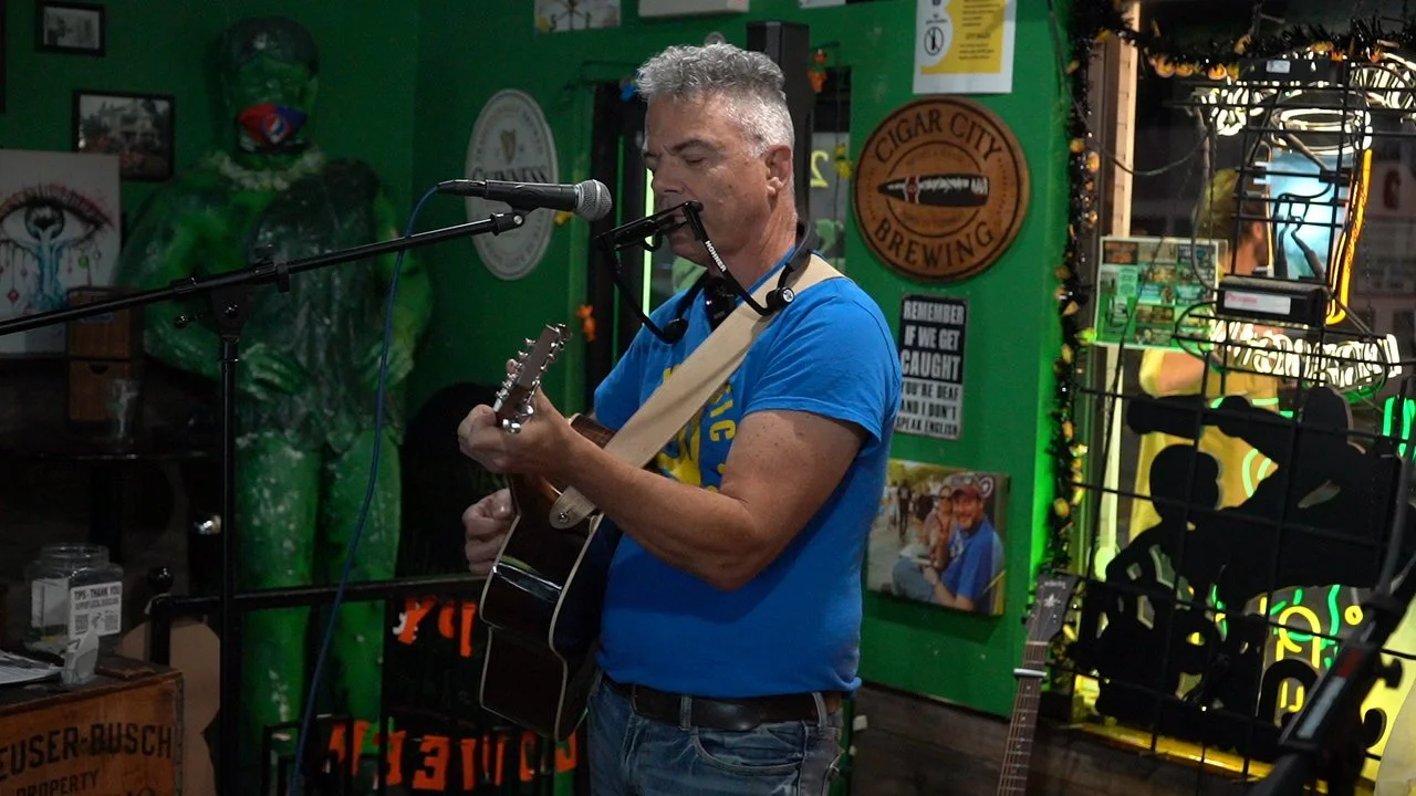 A man playing an acoustic guitar and singing into a microphone in a lively, decorated bar or pub with green walls, posters, and a large green mascot or statue in the background.