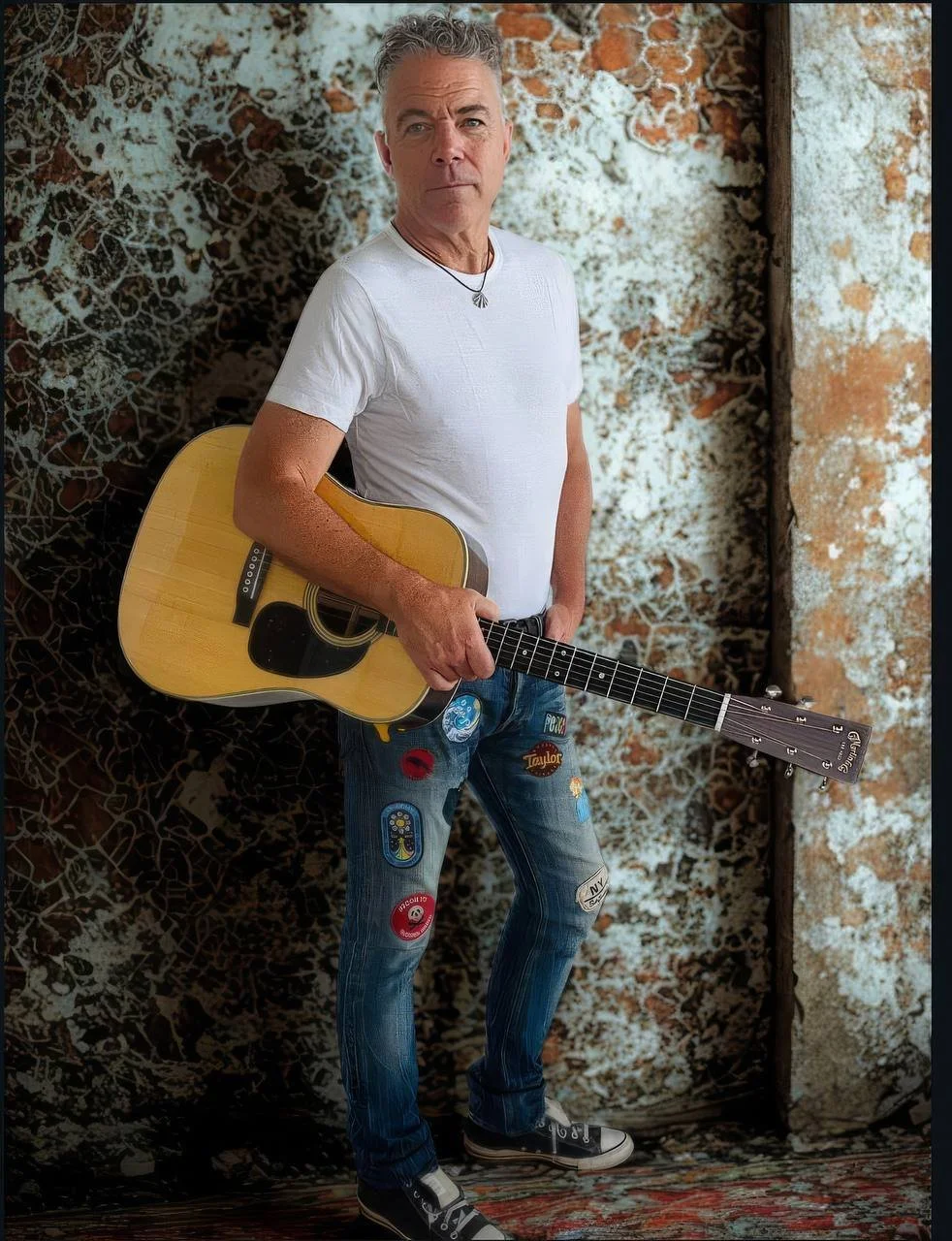 A man with gray hair wearing a white T-shirt and ripped jeans with patches, holding an acoustic guitar, standing in front of a textured, abstract background.