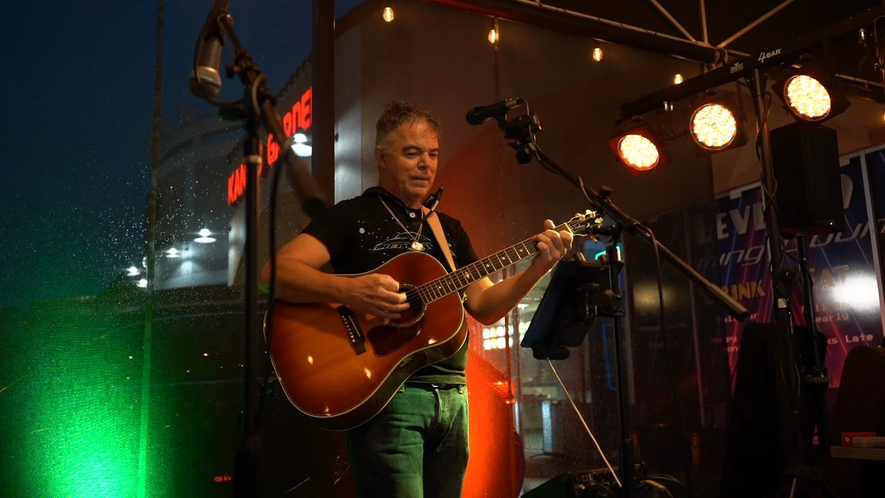Man playing acoustic guitar on stage at night with stage lights and a microphone, in a dimly lit venue.