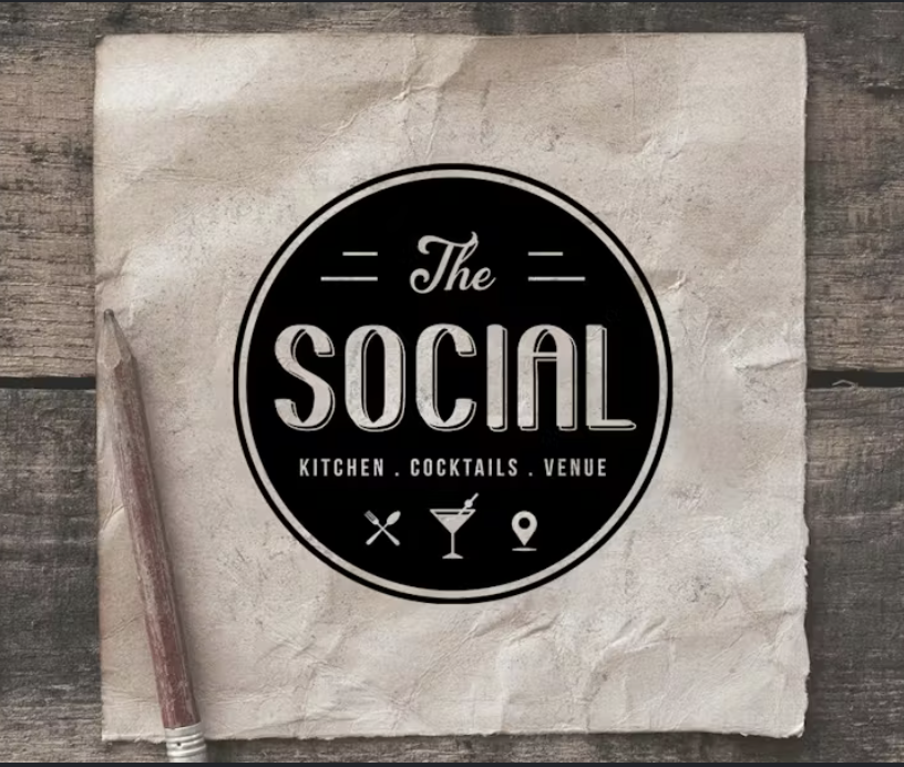 Logo for 'The Social' on crumpled paper, with icons of a fork, cocktail, and location pin, on a wooden surface.
