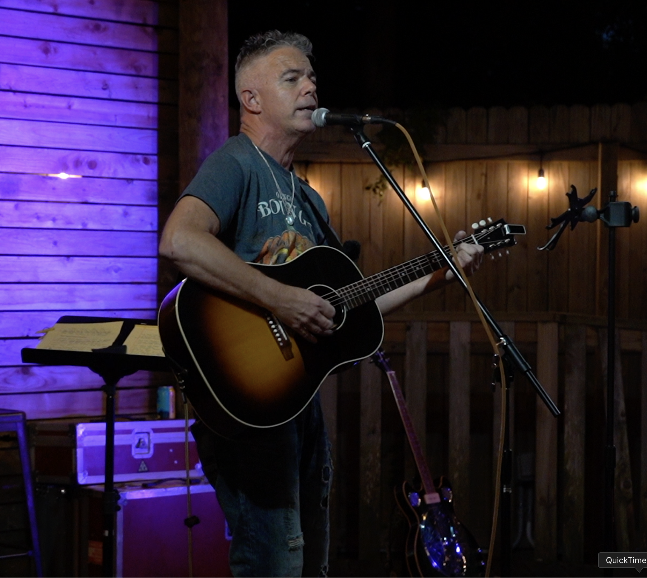 A man singing and playing an acoustic guitar on stage at a live music venue with wooden walls and warm lighting.