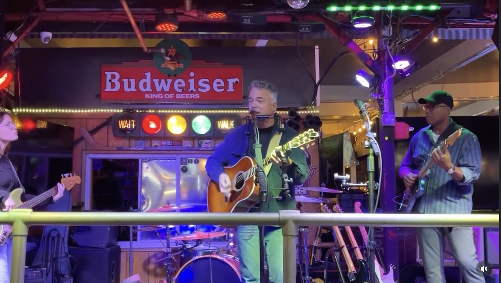 Musicians performing on stage at a bar with a Budweiser sign in the background, colorful lights, and musical instruments.