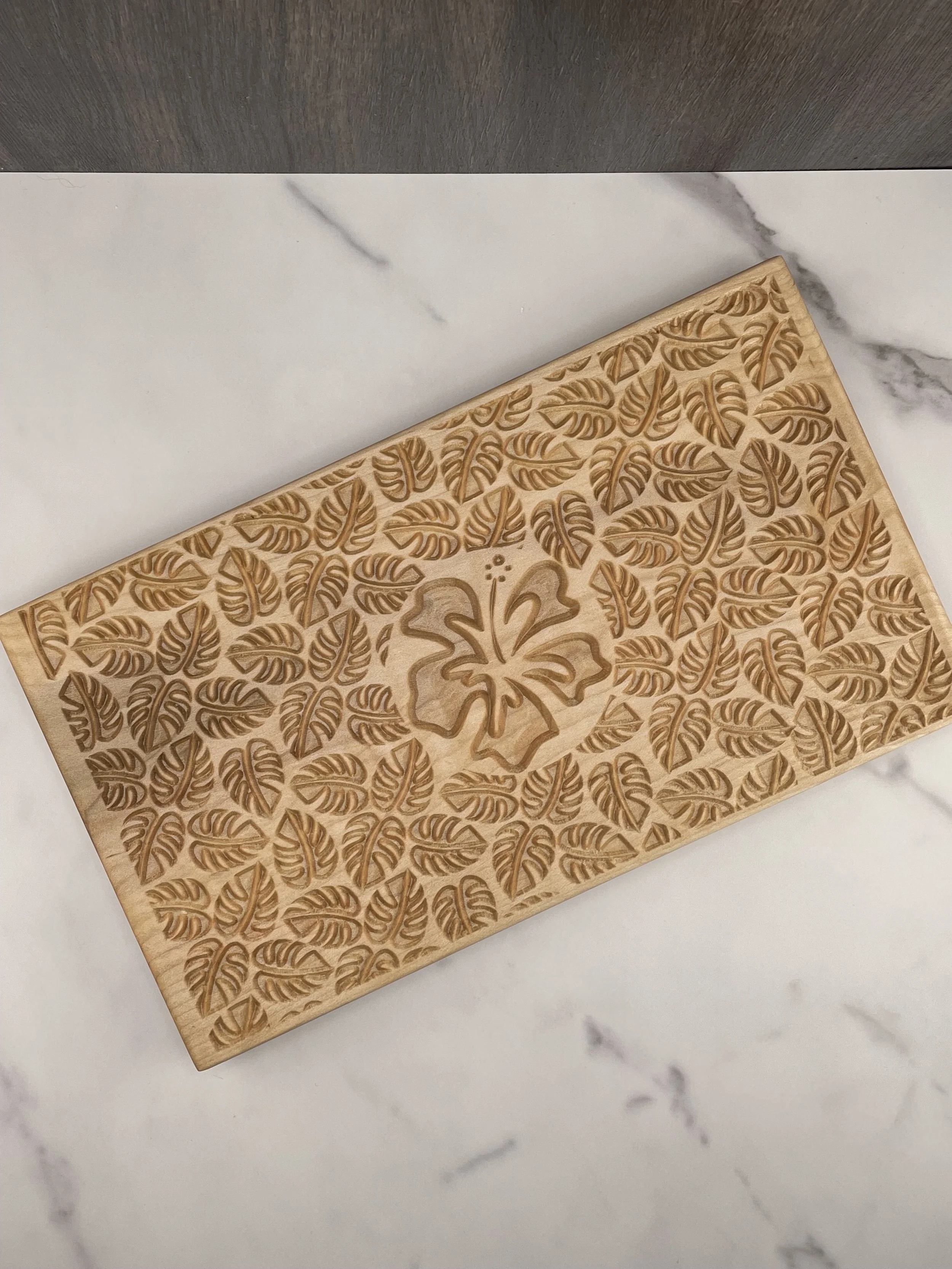 A wooden cutting board with a carved floral and leaf pattern, placed on a white marble surface.