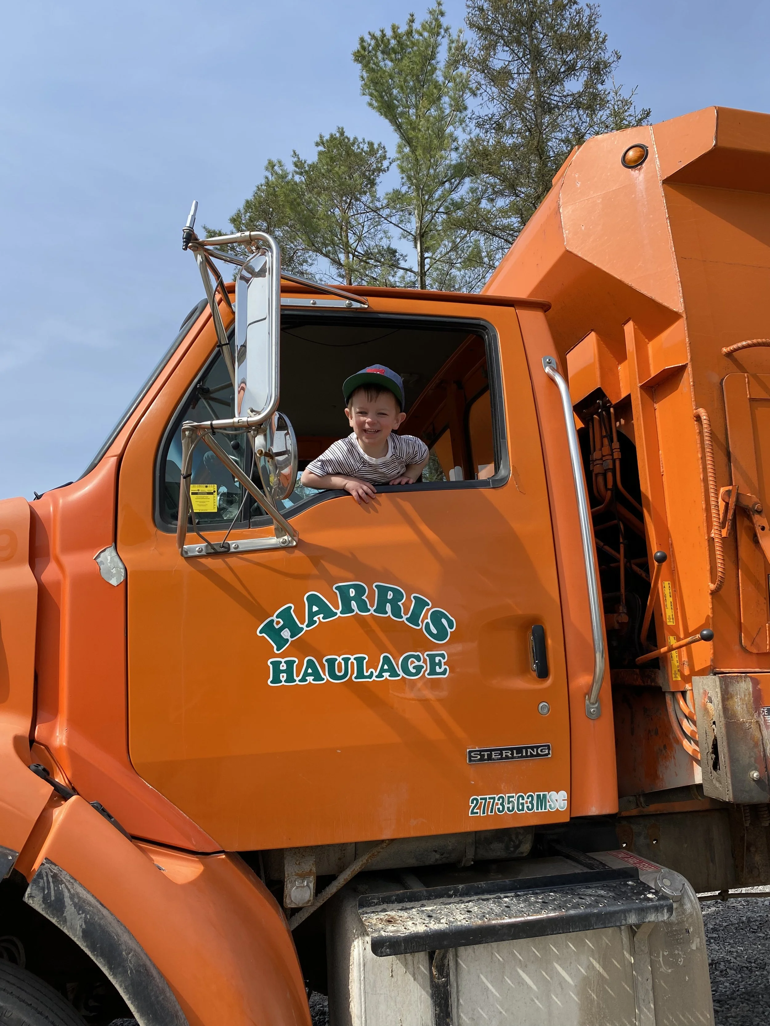 A young boy smiling and leaning out of the window of an orange Harris Haulage dump truck.