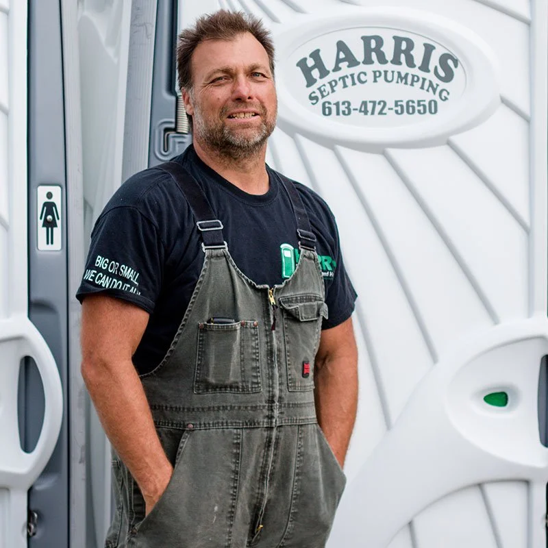A man with a beard and brown hair standing outdoors, wearing a black t-shirt and gray overalls, in front of a white portable restroom with a sign that reads 'Harris Septic Pumping' along with a phone number.