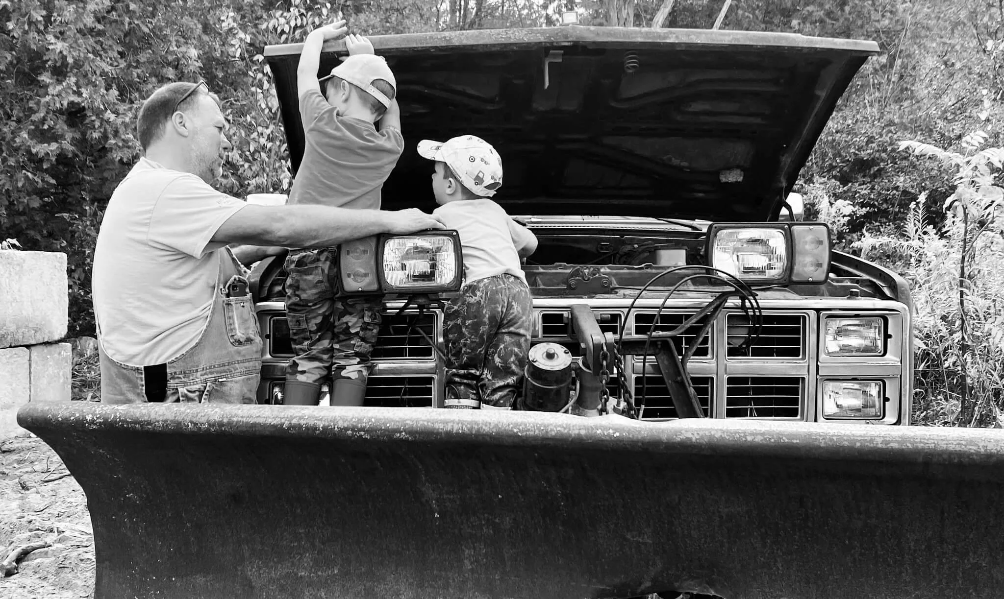 A man and two children working on a car engine outdoors, with the hood open, surrounded by trees and foliage.