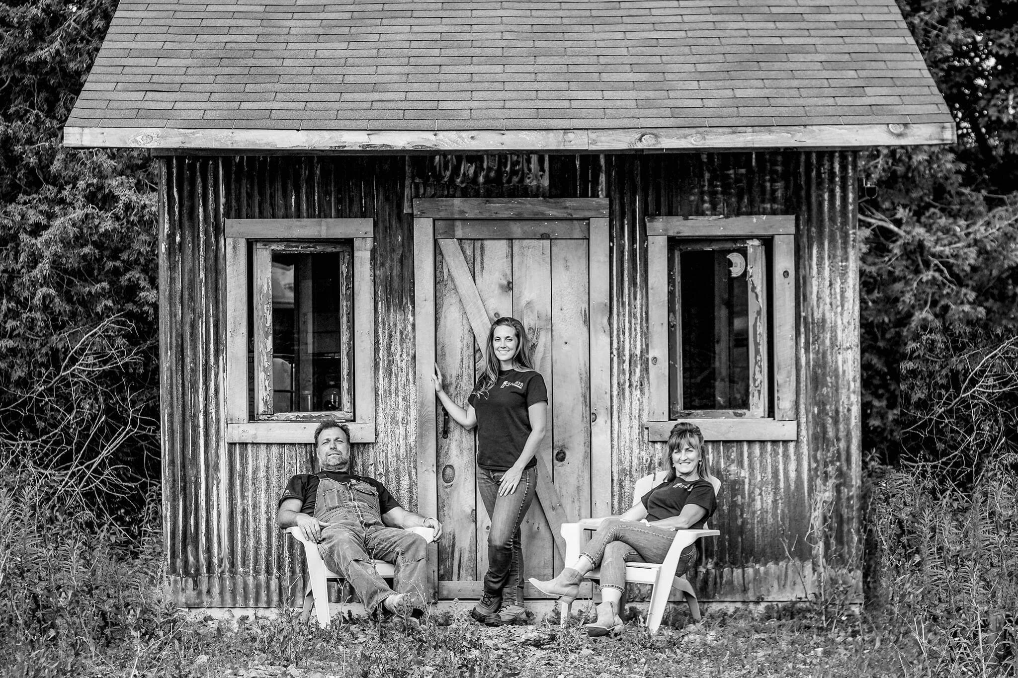 Three people sitting and standing in front of a rustic wooden barn with boarded-up windows, black and white photo.