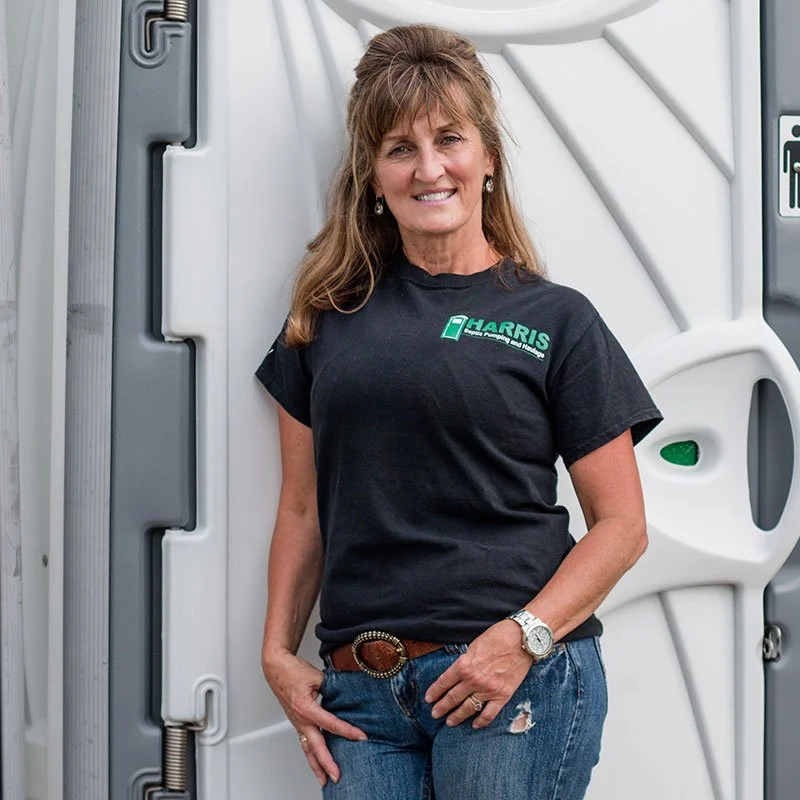 Woman smiling and standing in front of an RV, wearing a black T-shirt with green text and blue jeans.