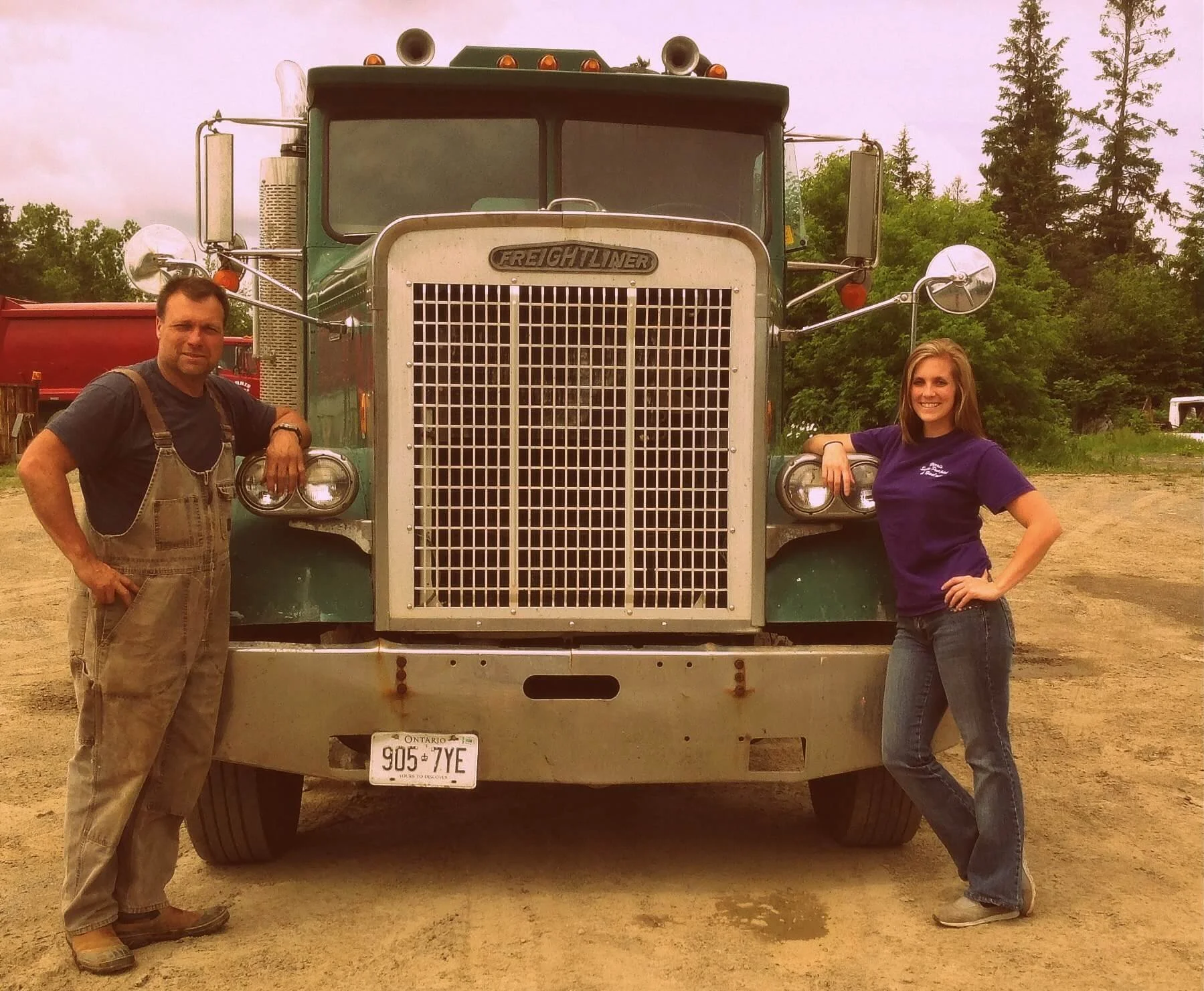 A man and a woman standing beside a large green Freightliner semi-truck with a chrome grille on a dirt lot, with trees in the background.