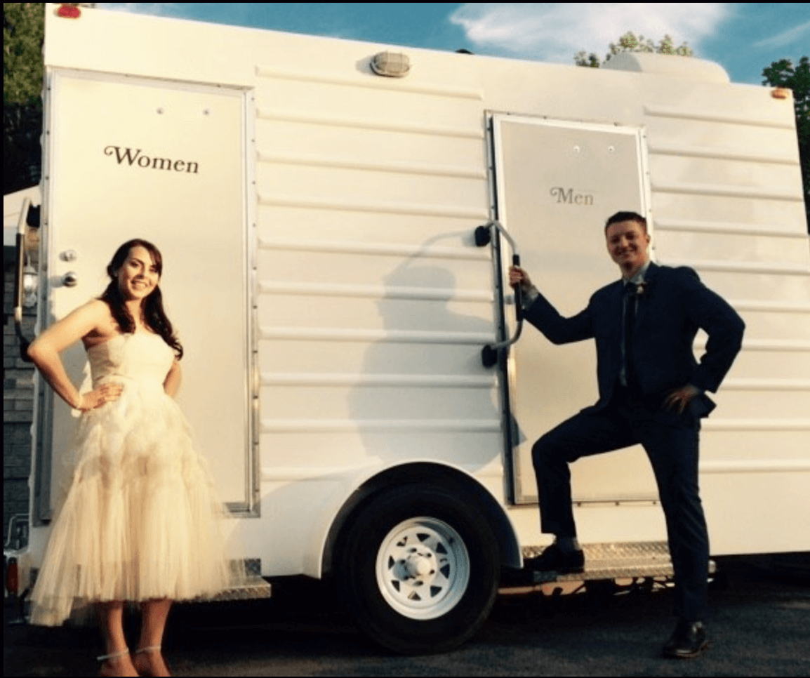 A woman in a beige dress and a man in a suit posing in front of a white portable restroom trailer labeled 'Women' and 'Men' during the daytime.