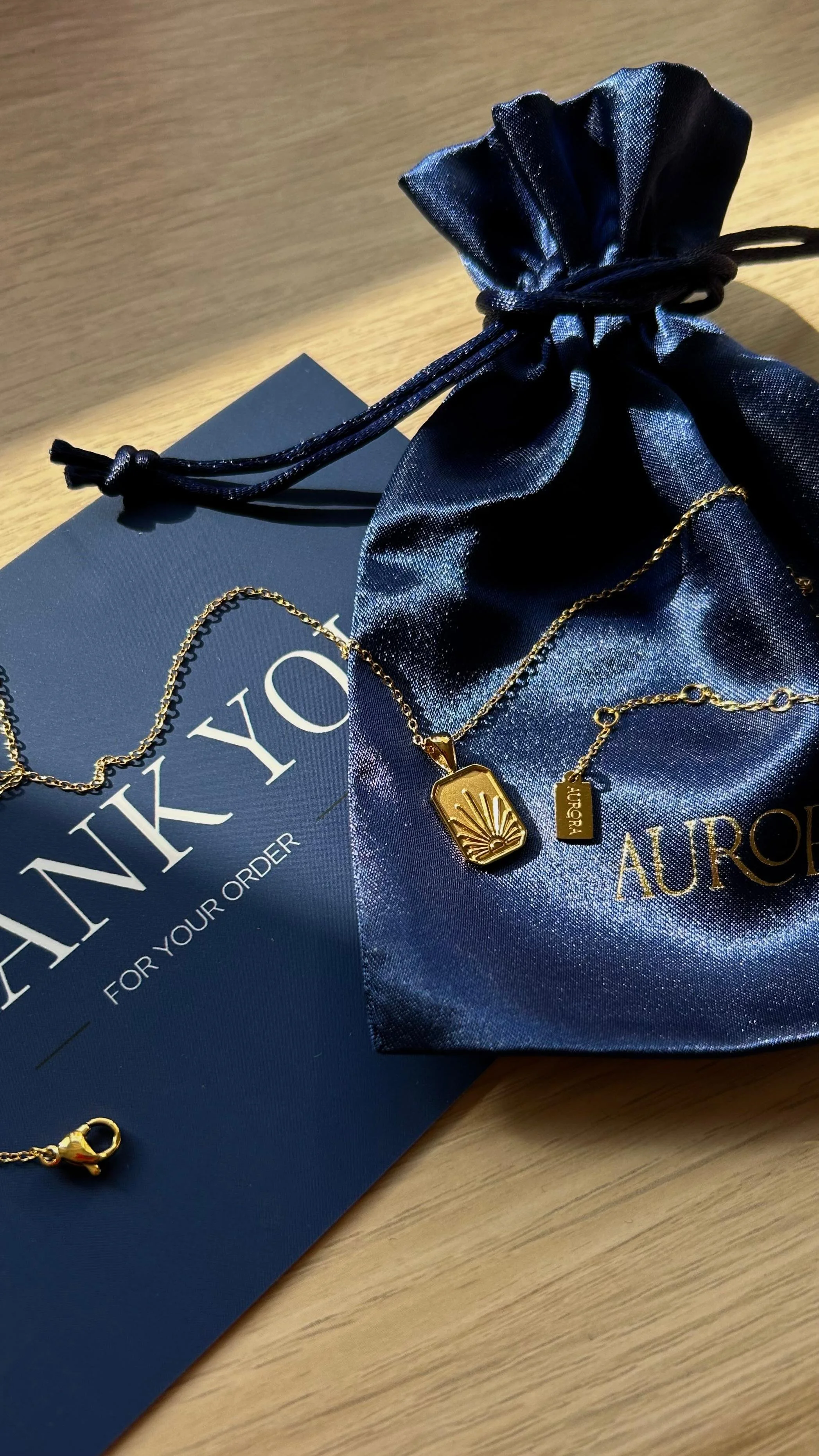 Gold jewelry necklaces and a bracelet on a navy blue jewelry pouch and a dark blue shopping bag from AURAI, on a wooden surface.