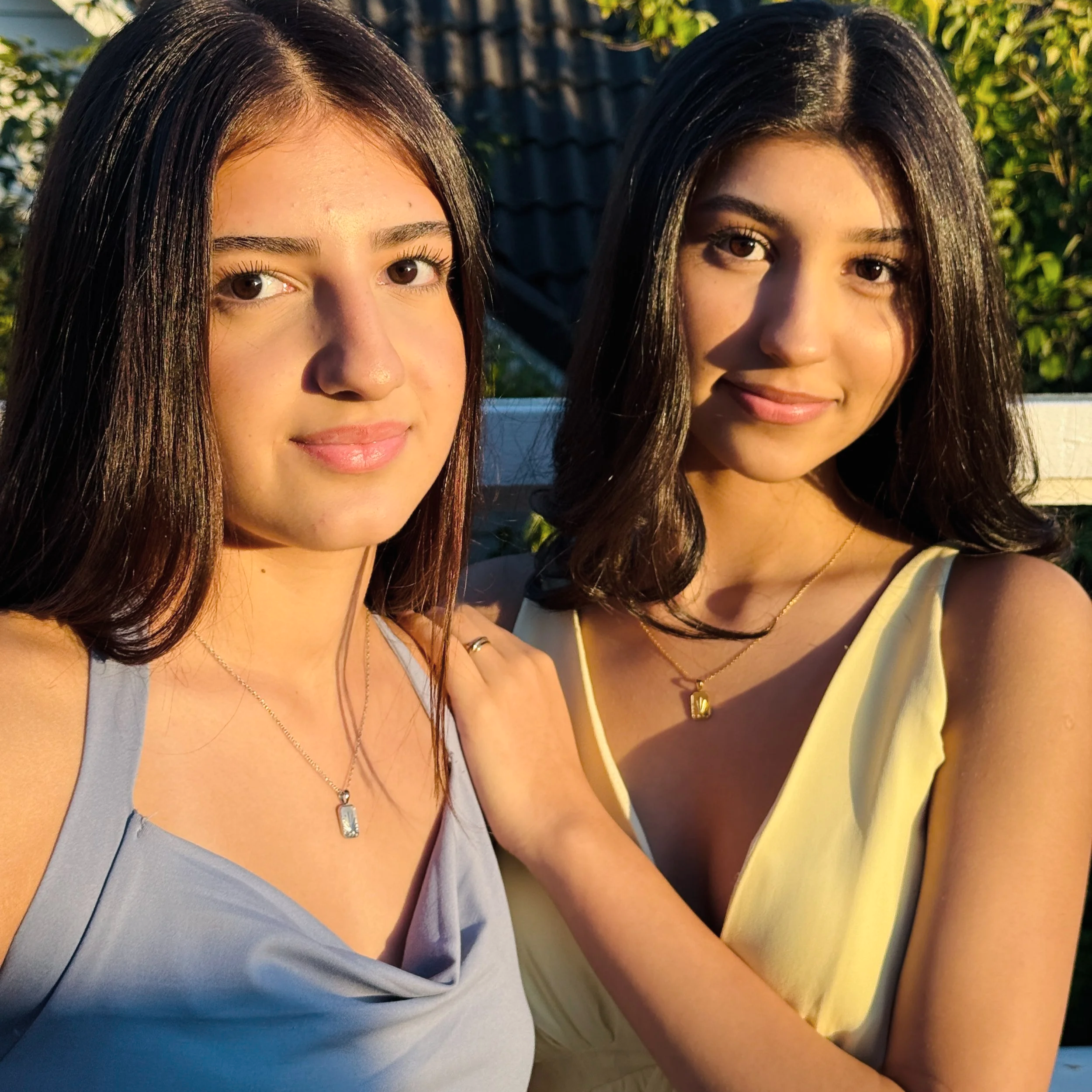 Two young women with dark hair and medium skin tones posing outdoors during golden hour. One women is wearing a light gray sleeveless top and the other a yellow sleeveless dress. They are smiling softly, and one has her arm around the other's shoulder. They wear necklaces with rectangular pendants.