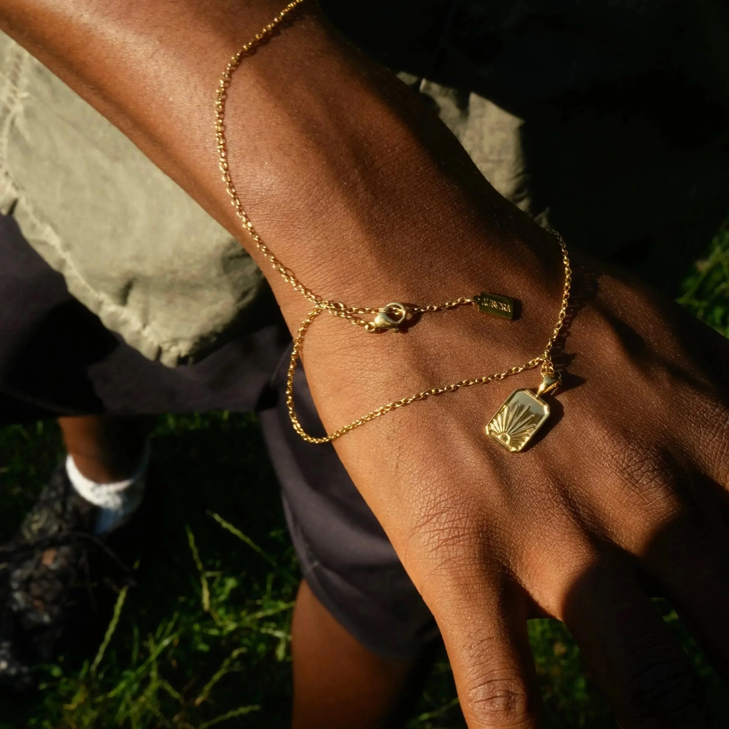 Close-up of a person's hand and wrist adorned with layered gold necklaces against a grassy background.