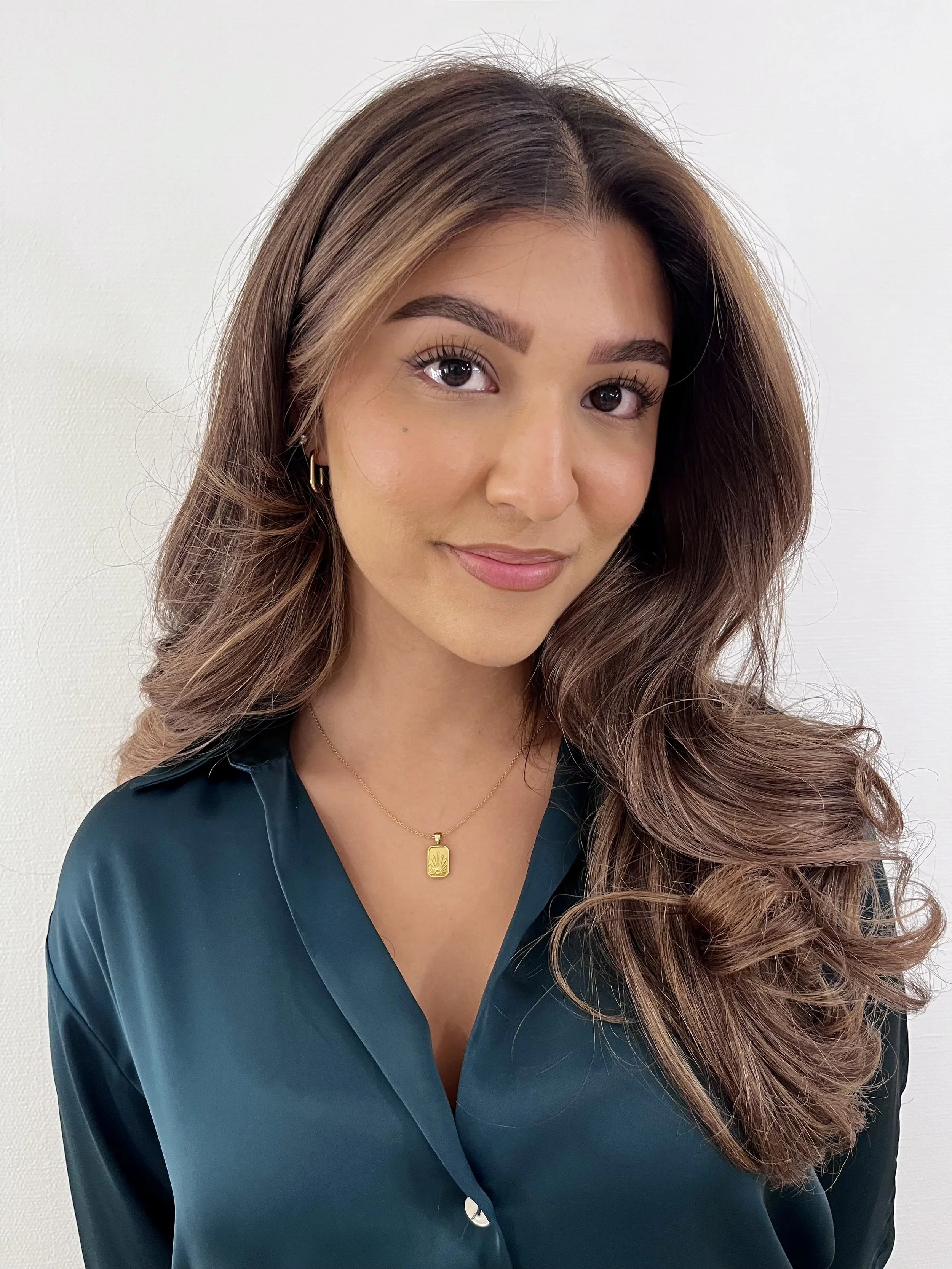 A woman with long, wavy brown hair wearing a dark teal satin blouse and a gold necklace with a rectangular pendant, standing against a plain white background.