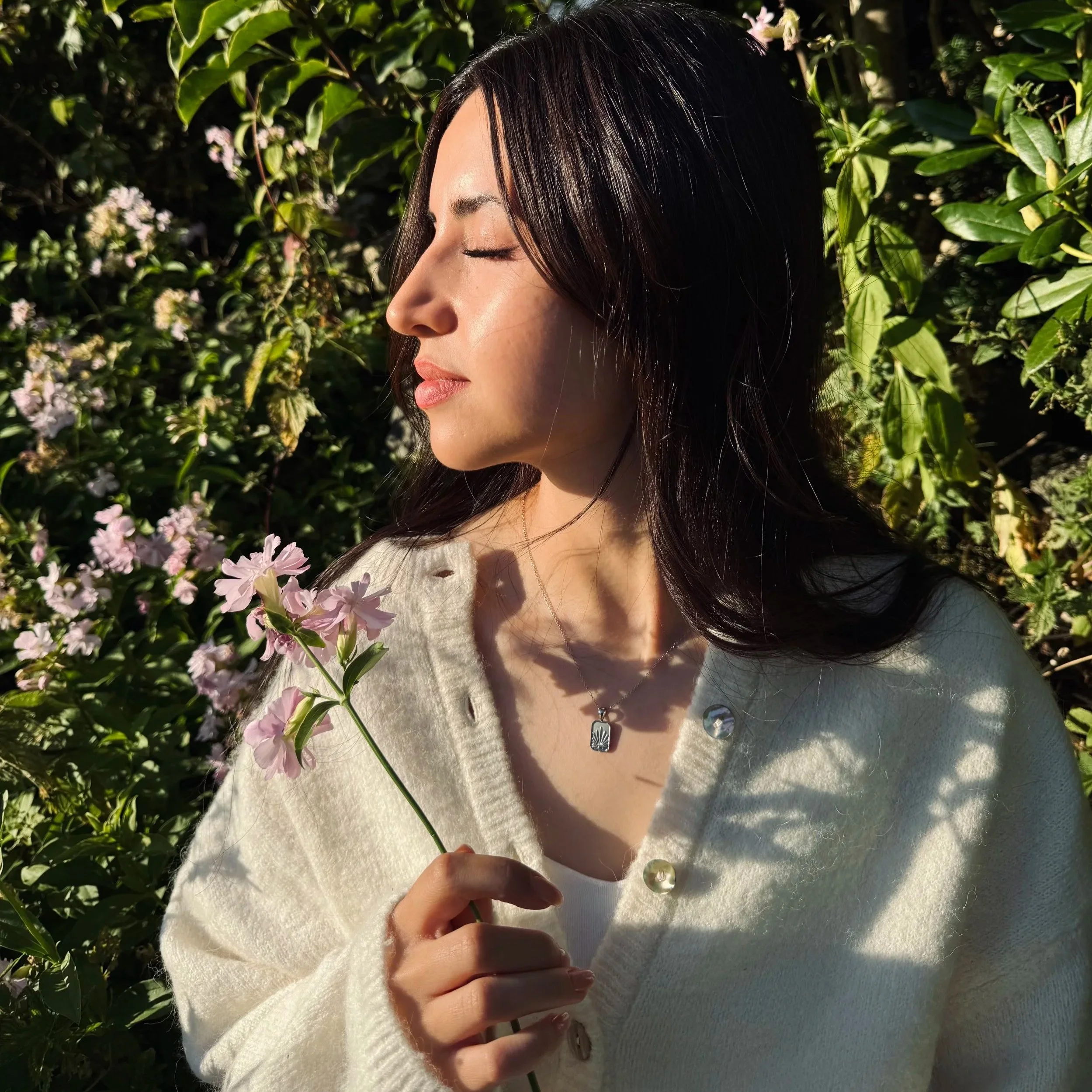A woman with closed eyes and dark hair stands among greenery, holding a pink flower near her chest, wearing a cream-colored cardigan and silver jewelry.