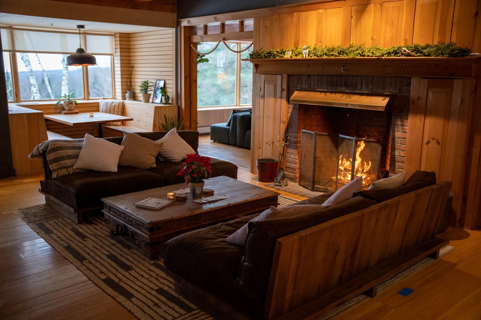Cozy living room with a stone fireplace, wood paneled walls, and a fire burning. Sofa with cushions, a wooden coffee table with a poinsettia, magazines, and candles. Large windows with a view of trees outside, creating a warm and inviting atmosphere.