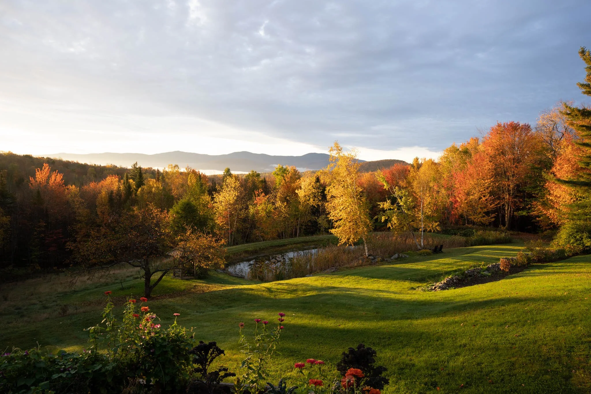 Scenic landscape of a lush green lawn with colorful fall trees, a small pond, and distant mountains under a partly cloudy sky during sunset.