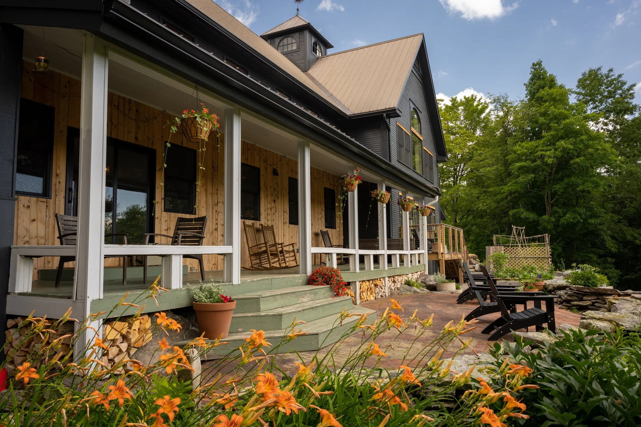 A house with a wooden porch and black siding, surrounded by greenery and flowers, with a brick patio and outdoor chairs.