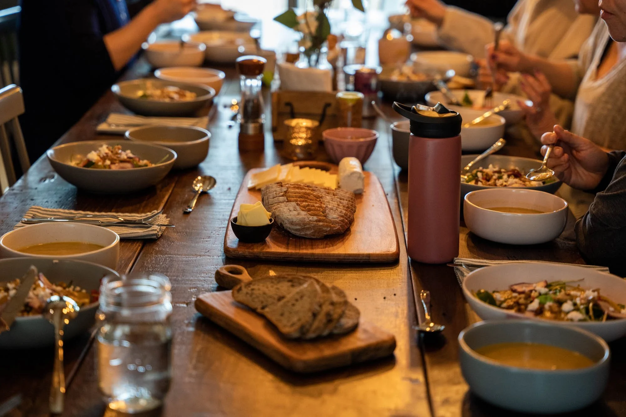 A long wooden dining table with various bowls of soup, slices of bread on a cutting board, a loaf of bread, and a pink water bottle. Several people are seated around the table, serving themselves and eating.