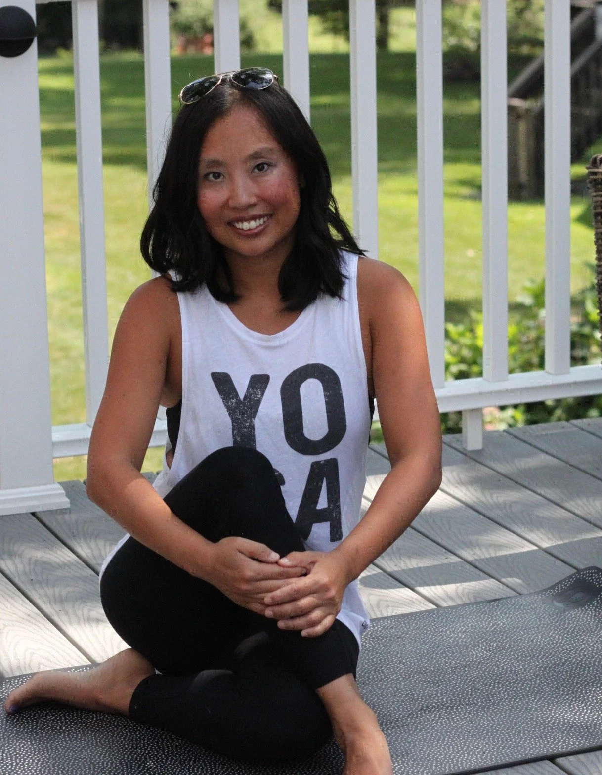 A woman with shoulder-length black hair, wearing sunglasses on her head and a sleeveless white tank top with black letters, sitting on a yoga mat on a wooden deck outdoors, with green grass and trees in the background.