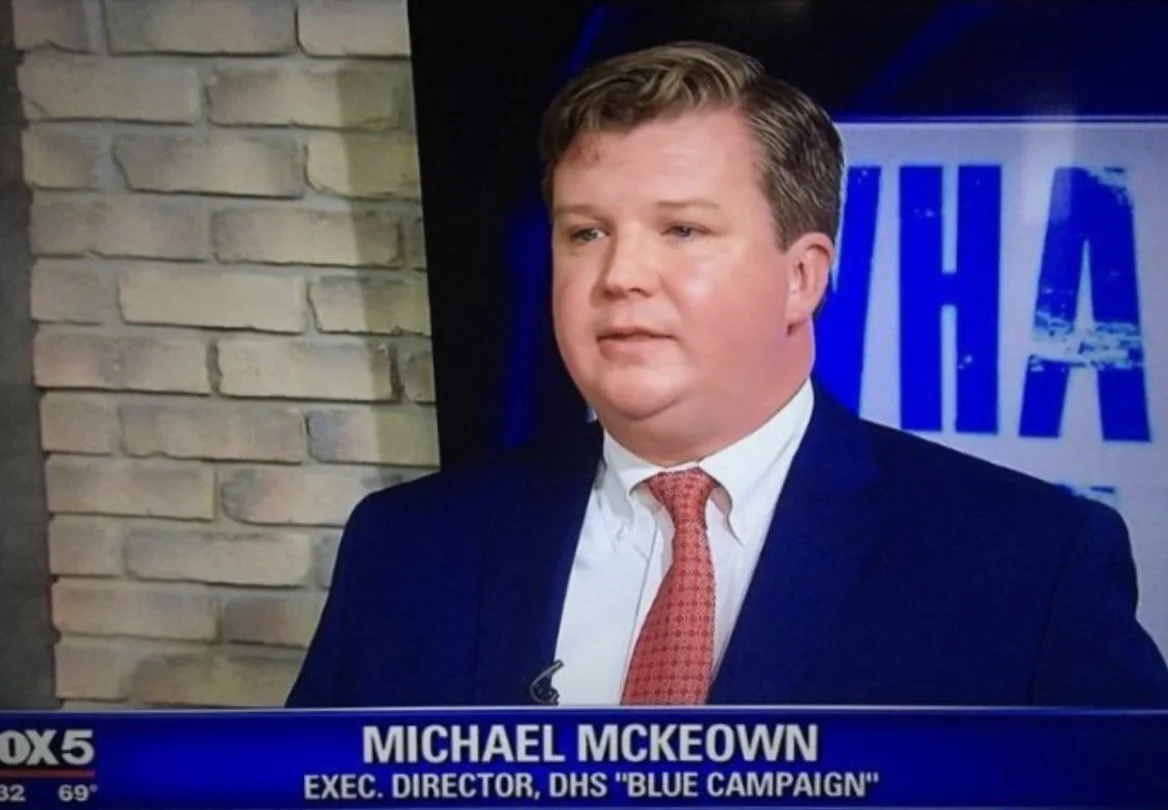 Television news interview with Michael McKeown, executive director of DHS 'Blue Campaign', in a studio with a brick wall background and a partially visible news logo.