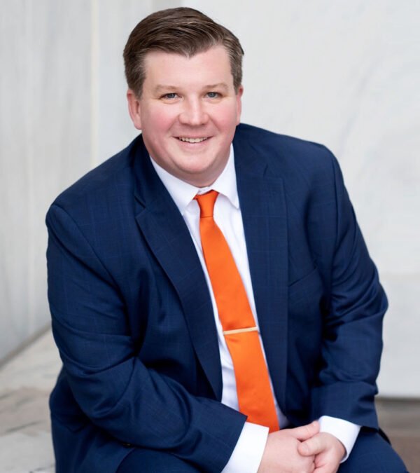 Portrait of a smiling man wearing a navy blue suit, white shirt, and orange tie, sitting indoors against a light-colored background.