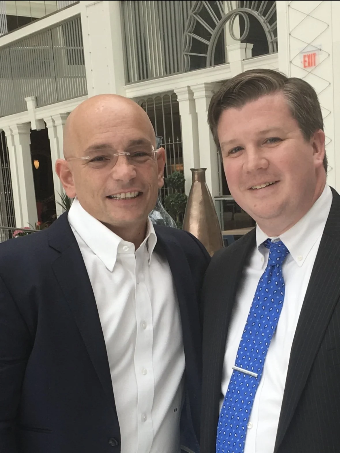 Two men in business suits smiling at the camera indoors, with decorative architecture and a large vase in the background.