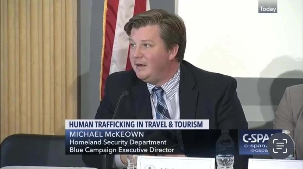 Michael McKeown speaking at a panel discussion on human trafficking in travel and tourism, with a water bottle and nameplate in front of him, an American flag behind, and a C-SPAN logo in the bottom right corner.