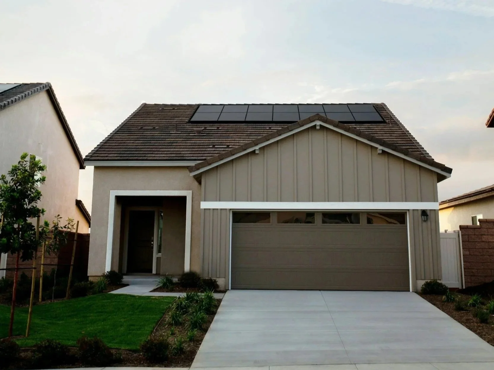Front view of a modern house with a beige roof and solar panels, beige and white exterior, garage door, small front yard with grass and plants, driveway, and neighboring houses visible.