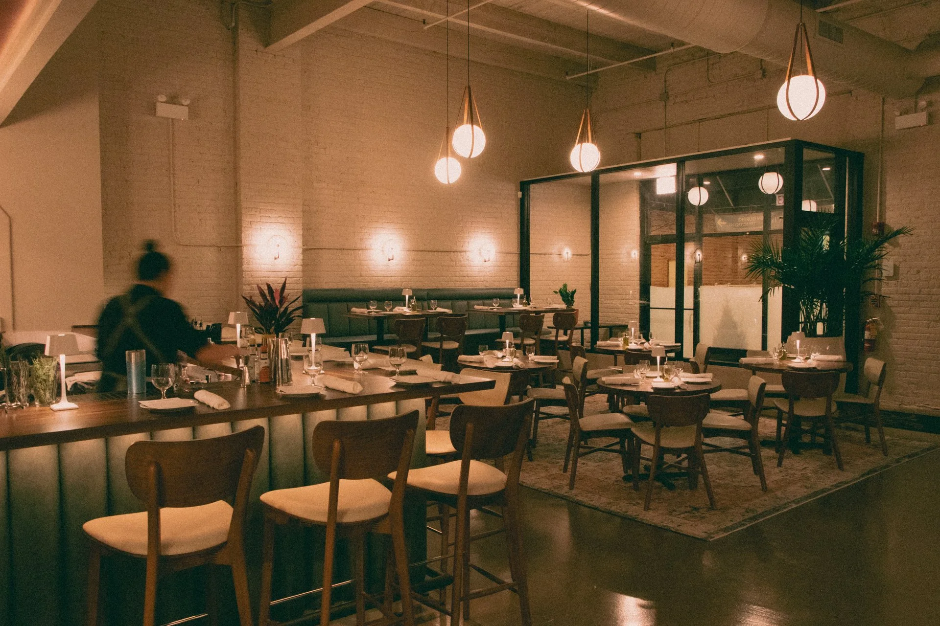 A restaurant bar and lounge with warm lighting, wooden furniture, white table settings, and potted plants, with a blurred figure of a person preparing a drink at the bar.