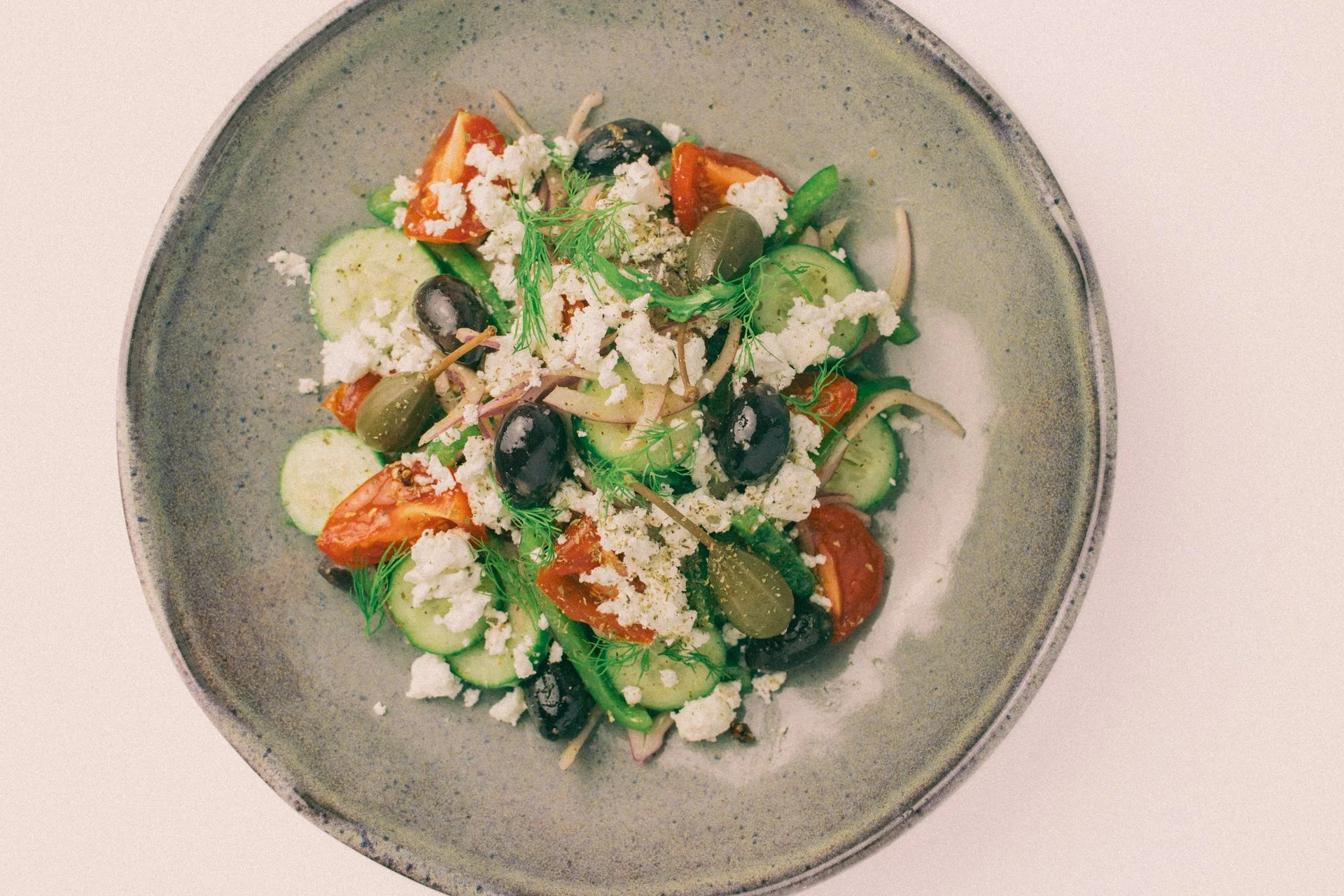 A bowl of Greek salad with cucumbers, cherry tomatoes, black olives, onion, feta cheese, and fresh herbs.