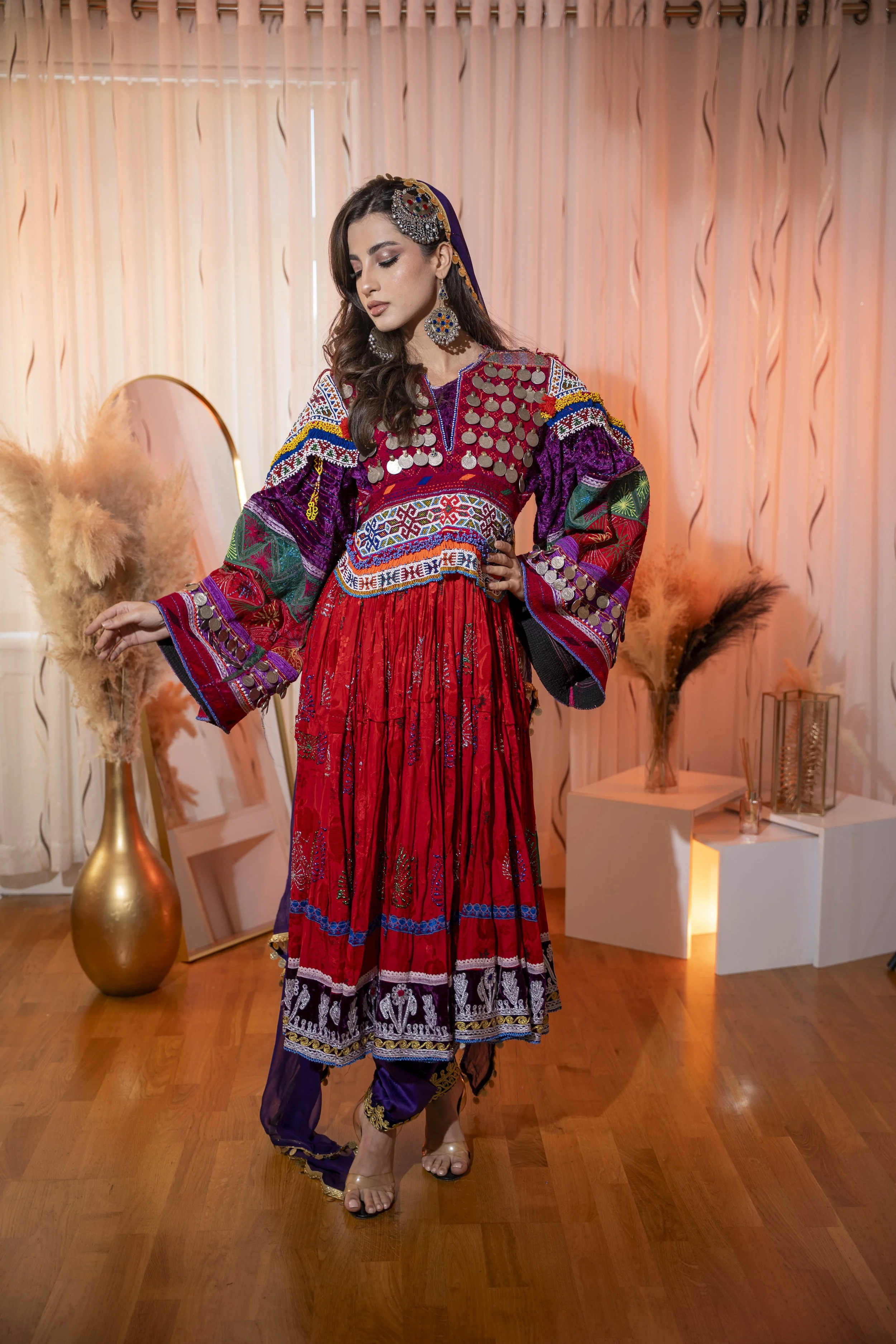 A woman in a colorful traditional Afghan dress with intricate embroidery and silver coin embellishments, standing indoors with a curtain backdrop, decorative vases with dried plants, and a mirror on the floor.
