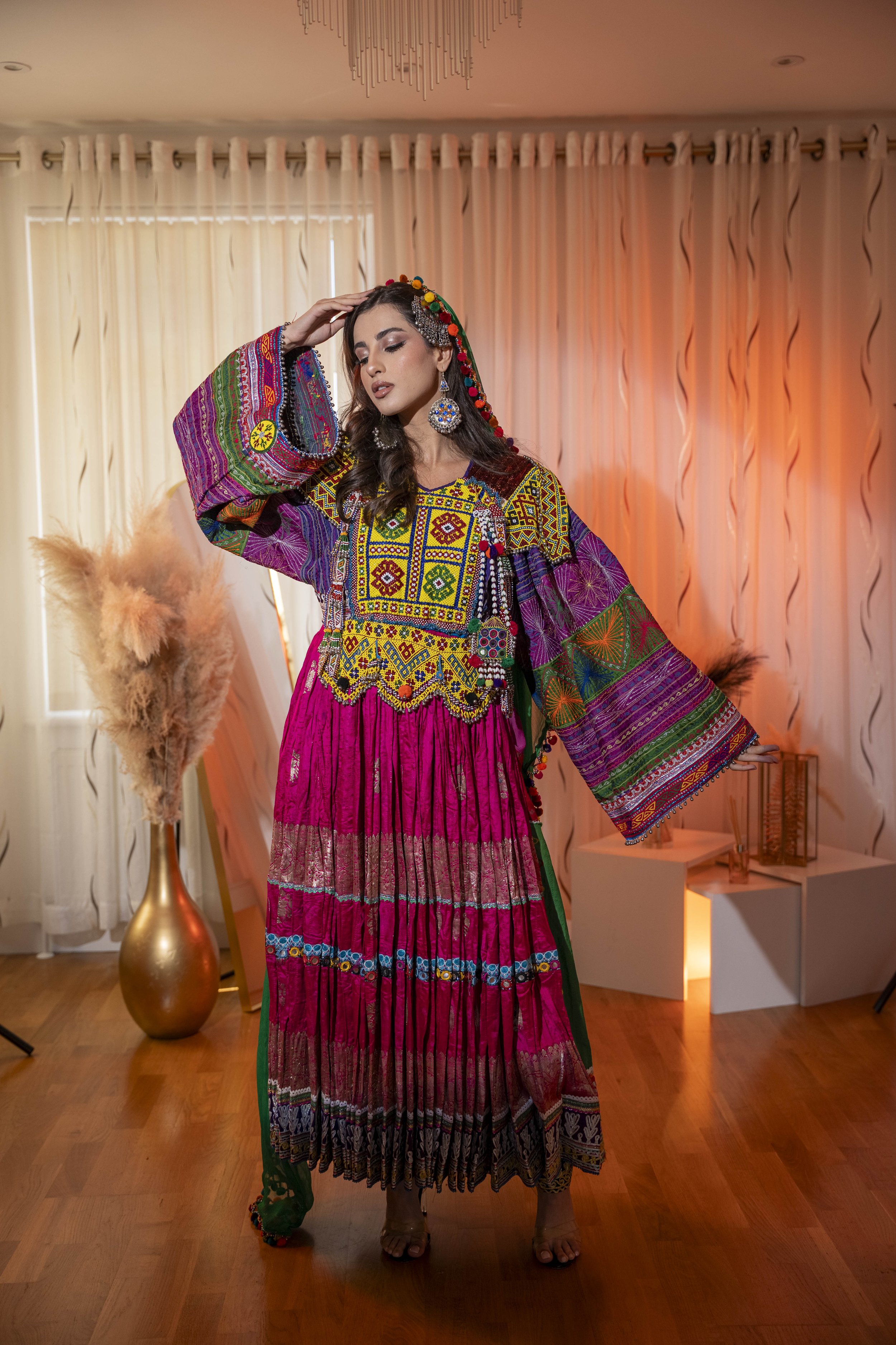 Woman wearing colorful traditional Afghan ethnic dress with intricate embroidery, standing in a room with wooden floors and beige curtains, posing with one hand on her head.