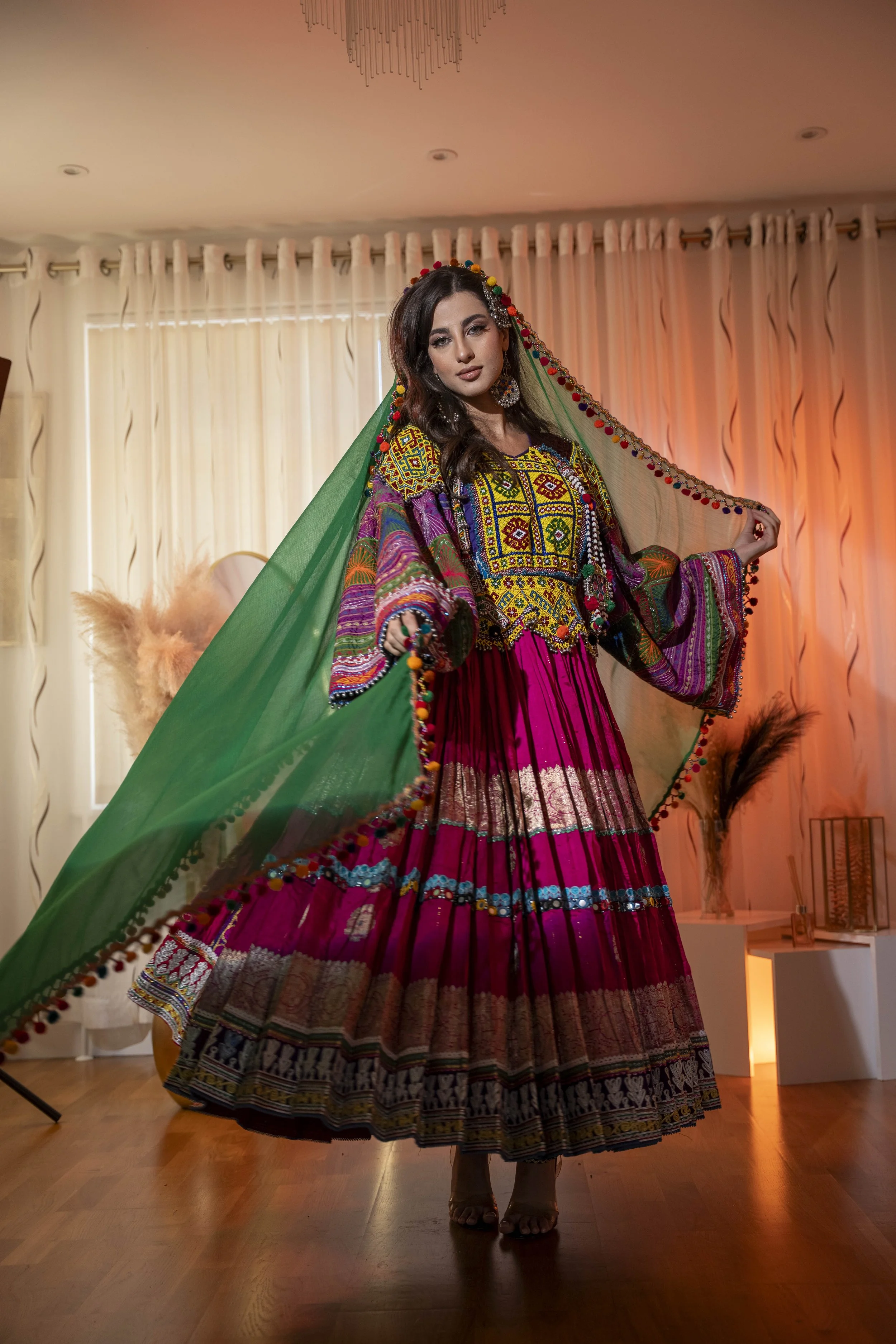 A woman wearing a traditional, colorful, embroidered Afghan ethnic dress with a matching veil, standing in a warmly lit room with curtains and decorative plants.