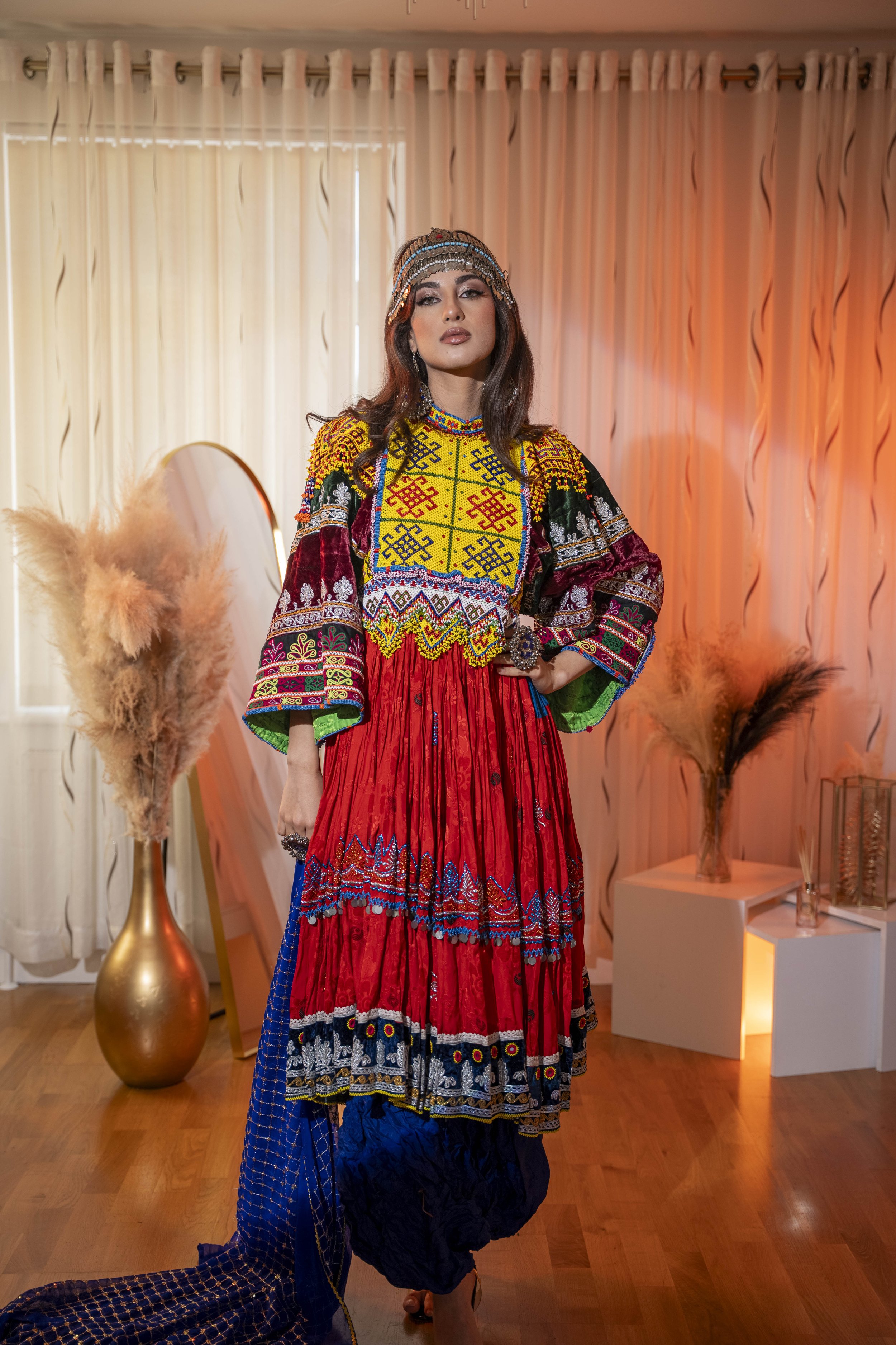 A woman in vibrant traditional ethnic Afghan attire standing in a room with artistic decor, including curtains, pampas grass in vases, and warm lighting.