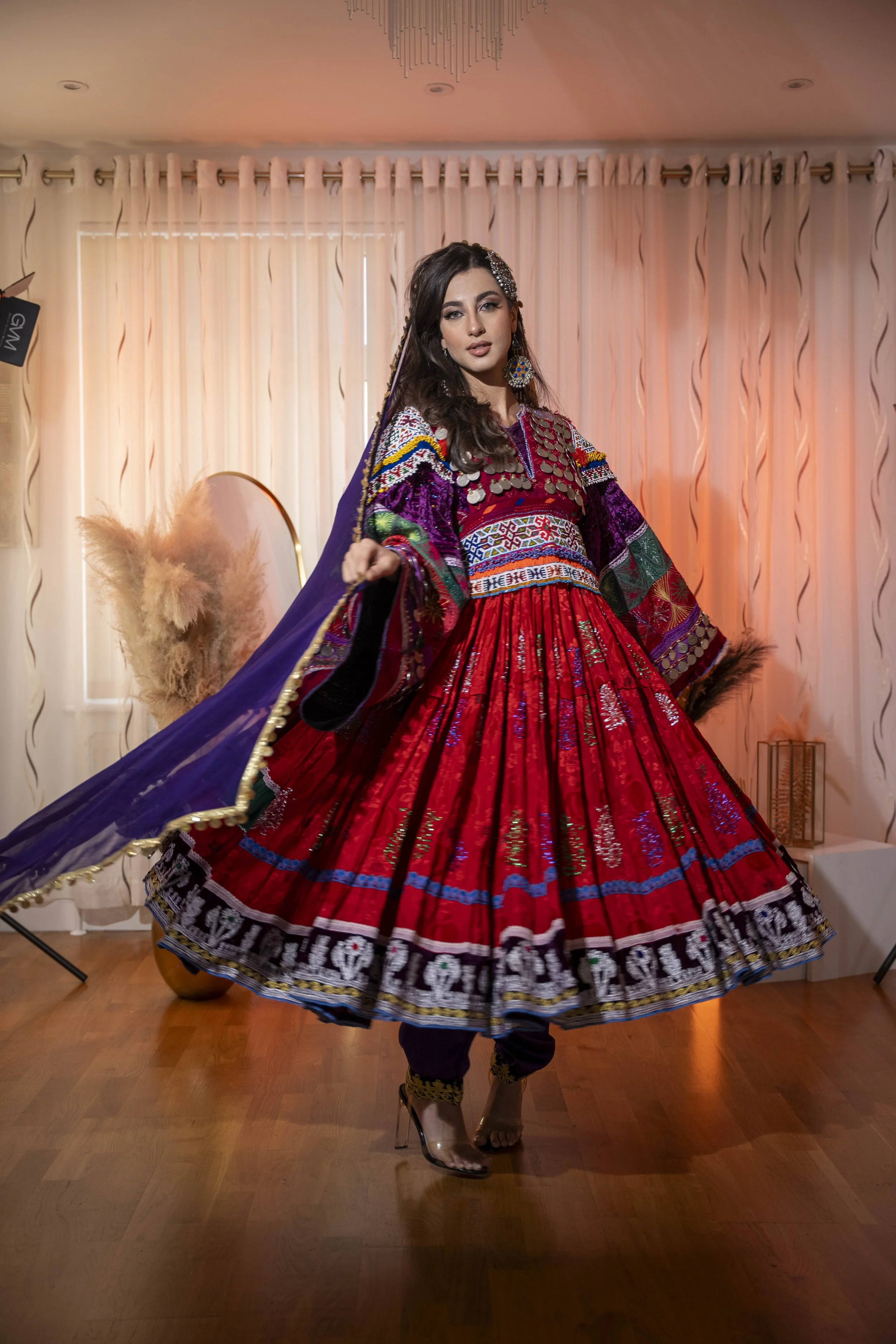 A woman wearing a vibrant, traditional, colorful Afghan dress with intricate embroidery, standing indoors with a backdrop of light-colored curtains, a modern lamp, and decorative plants.