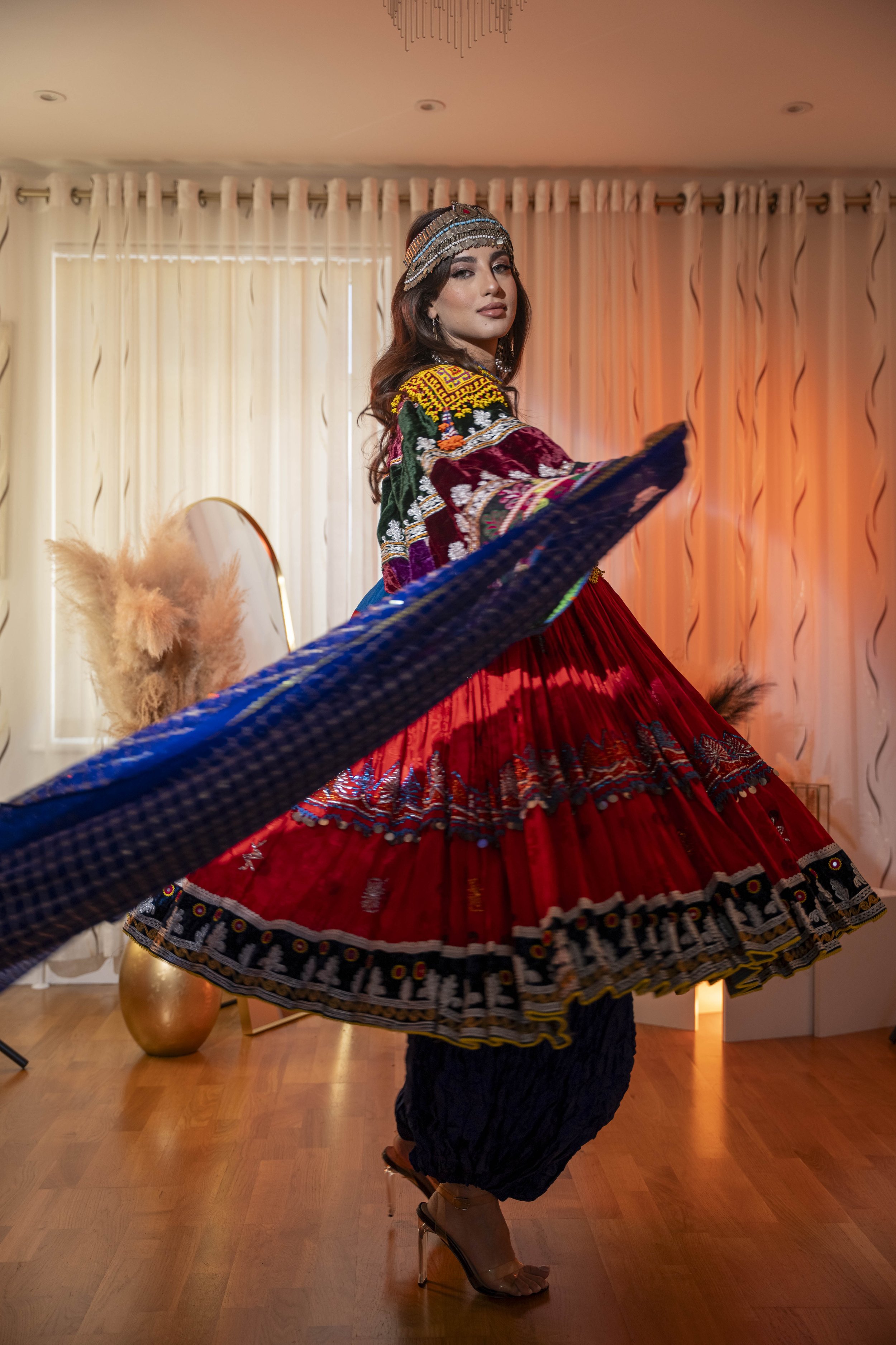 A woman performing a traditional dance in colorful ethnic Afghan clothing attire in a well-lit room with curtains and decorative elements.