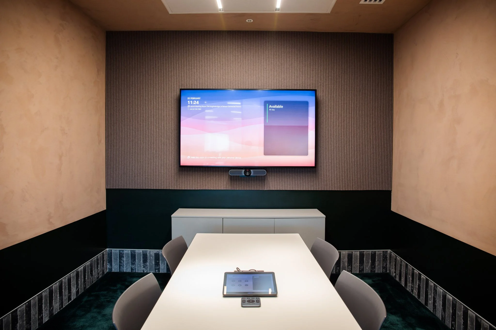 A modern conference room with a white table, four chairs, a large screen displaying the date and time, and a tablet on the table.