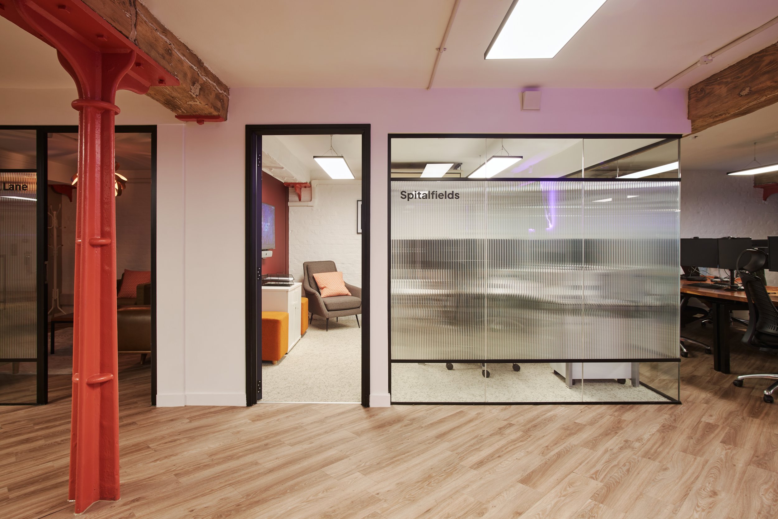 Office interior space with glass walls, wooden beams, and modern lighting. There is a waiting area with a chair and cushions, and a sign that reads 'Spitalfields' is visible on one of the glass partitions.