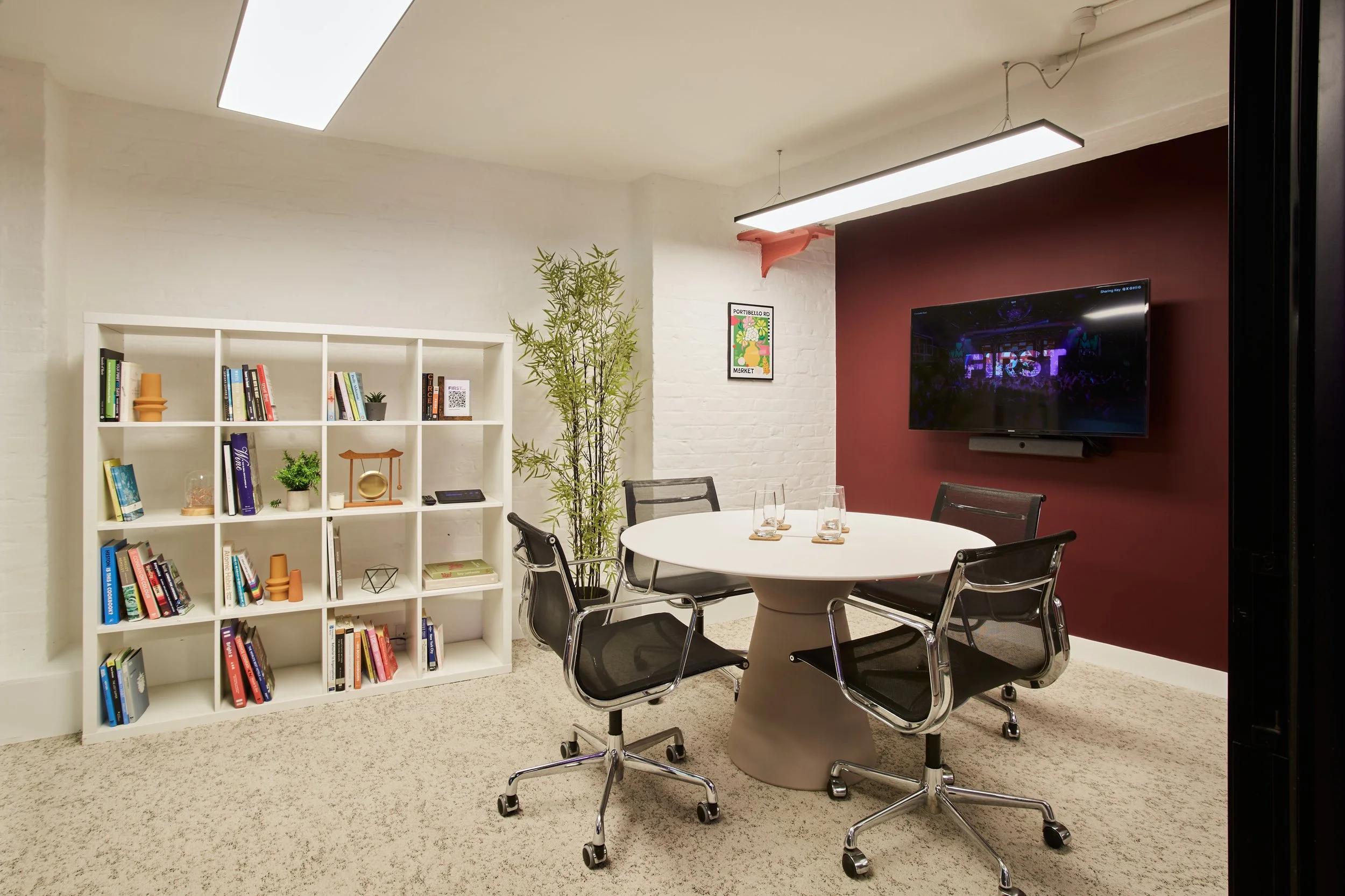 Modern conference room with white brick wall, a white bookshelf filled with books and decorative items, a large flat-screen TV on a maroon accent wall displaying the word "FIRST," a round table with four glasses and black office chairs, a tall potted