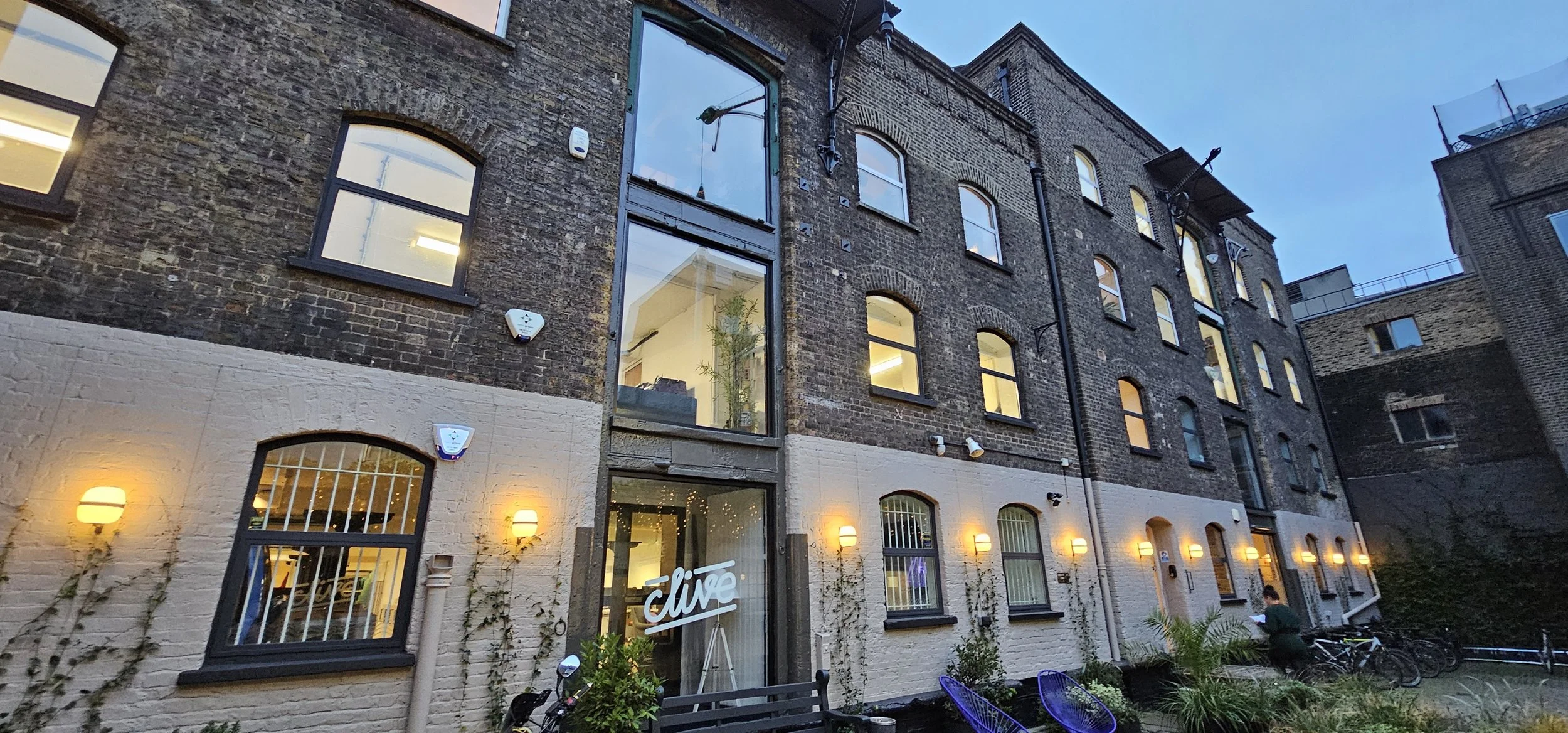 An exterior view of a multi-story brick building with large windows, some illuminated from inside. The building features a combination of brick and white-painted lower walls, with outdoor lighting, plants, and bicycles outside. It appears to be eveni