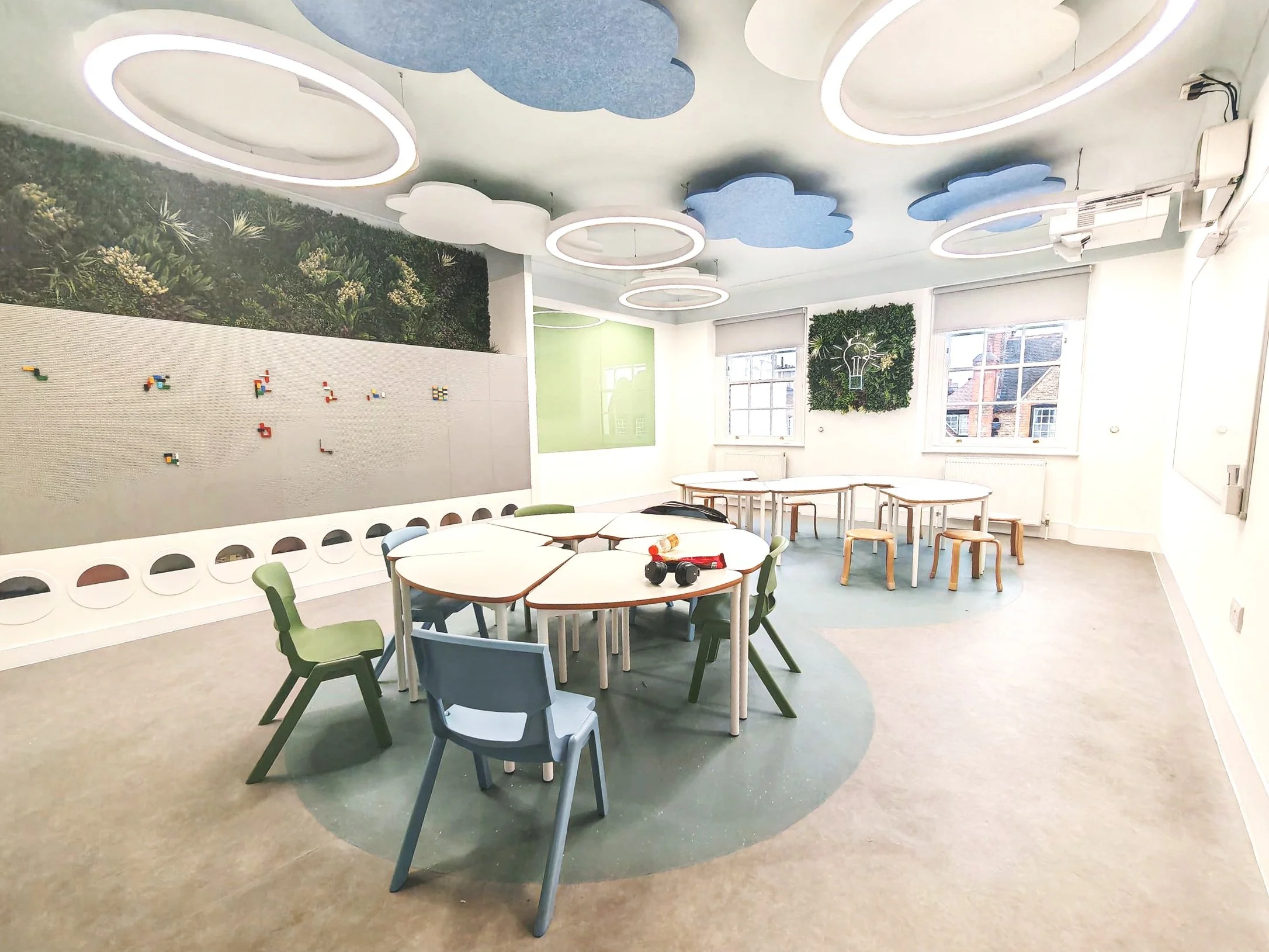 A bright, modern room with cloud-shaped and circular ceiling lights, a green and grassy wall with a light bulb design, large windows, and a circular arrangement of tables and chairs, with some small objects on the tables.