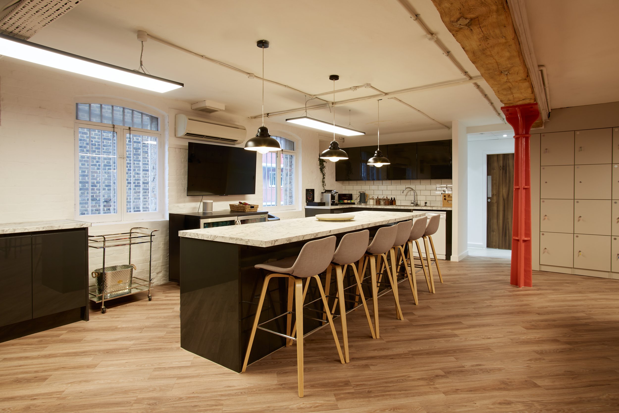 Interior of a modern kitchen with a large island, light-colored chairs, pendant lights, a wall-mounted TV, white brick backsplash, and large windows with brick buildings outside.