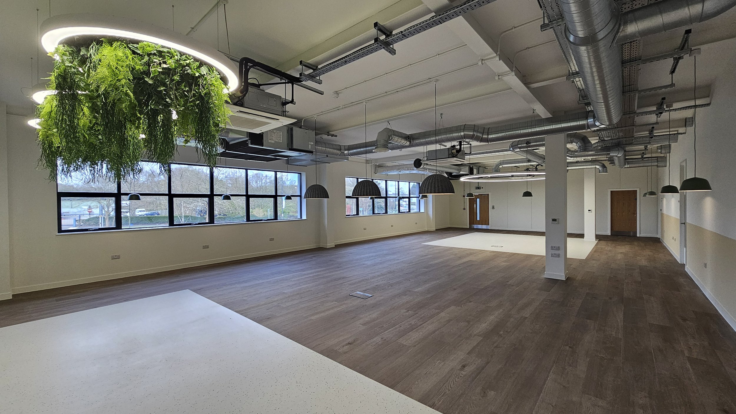 Empty modern office or studio with large windows, hanging pendant lights, ceiling ductwork, and a seating or workspace area with wood flooring and a white wall.