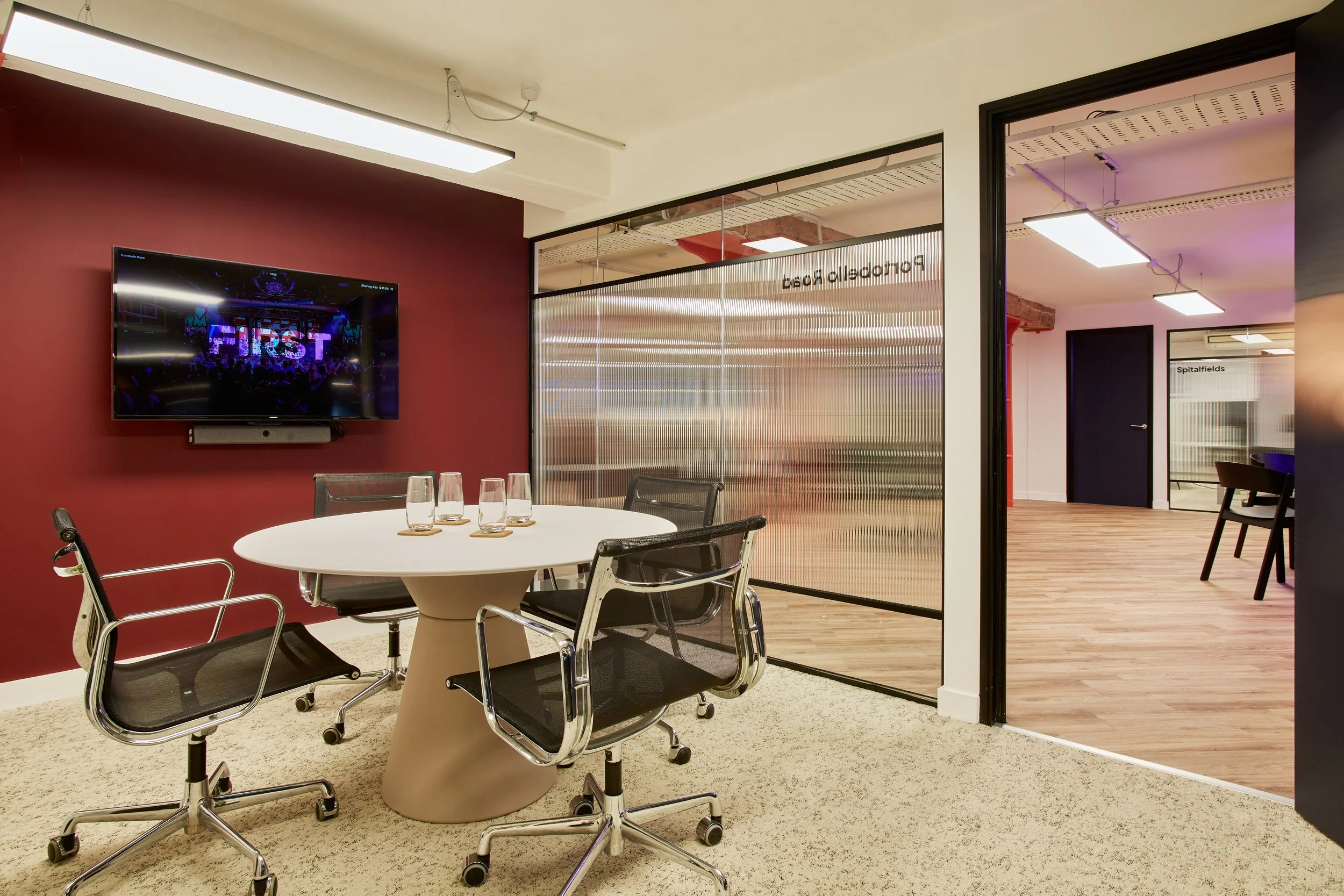 Modern conference room with round table, five glasses, black chairs, mounted TV showing a stage with the word "FIRST," glass partitions, and a doorway to another room.