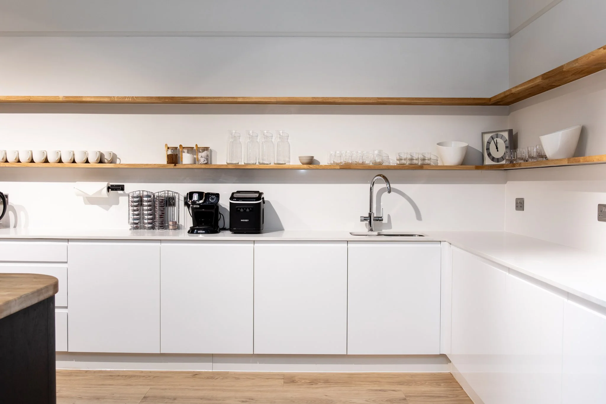 A modern kitchen with white cabinets, wooden open shelves, a sink, and various kitchen items including cups, glasses, jars, and a clock.